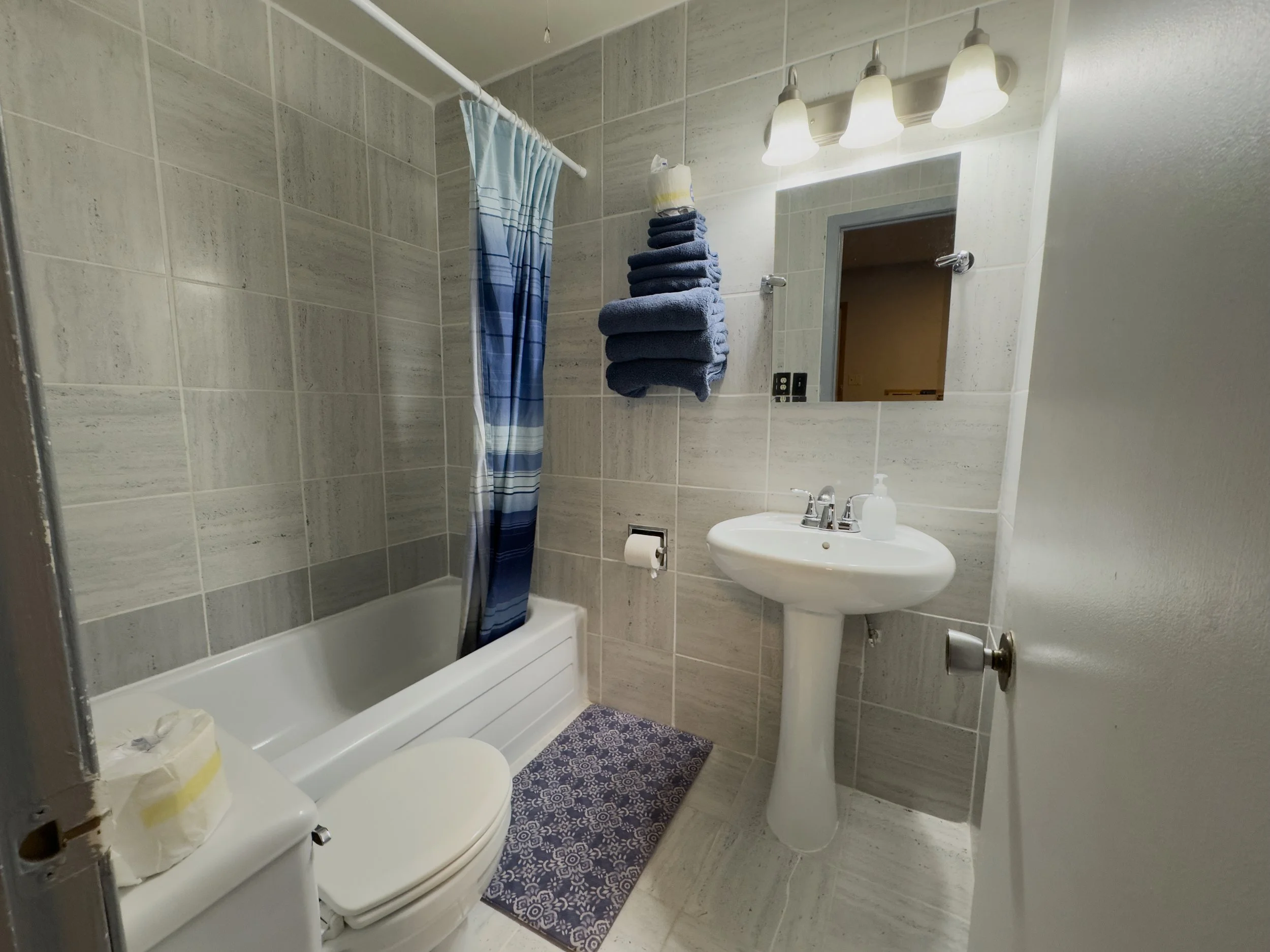 Small bathroom with beige tiled walls and white fixtures, including a toilet, pedestal sink with soap dispenser, mirror with three light fixtures above, a window with a blue gradient shower curtain, and a pile of folded dark blue towels on a wall shelf.
