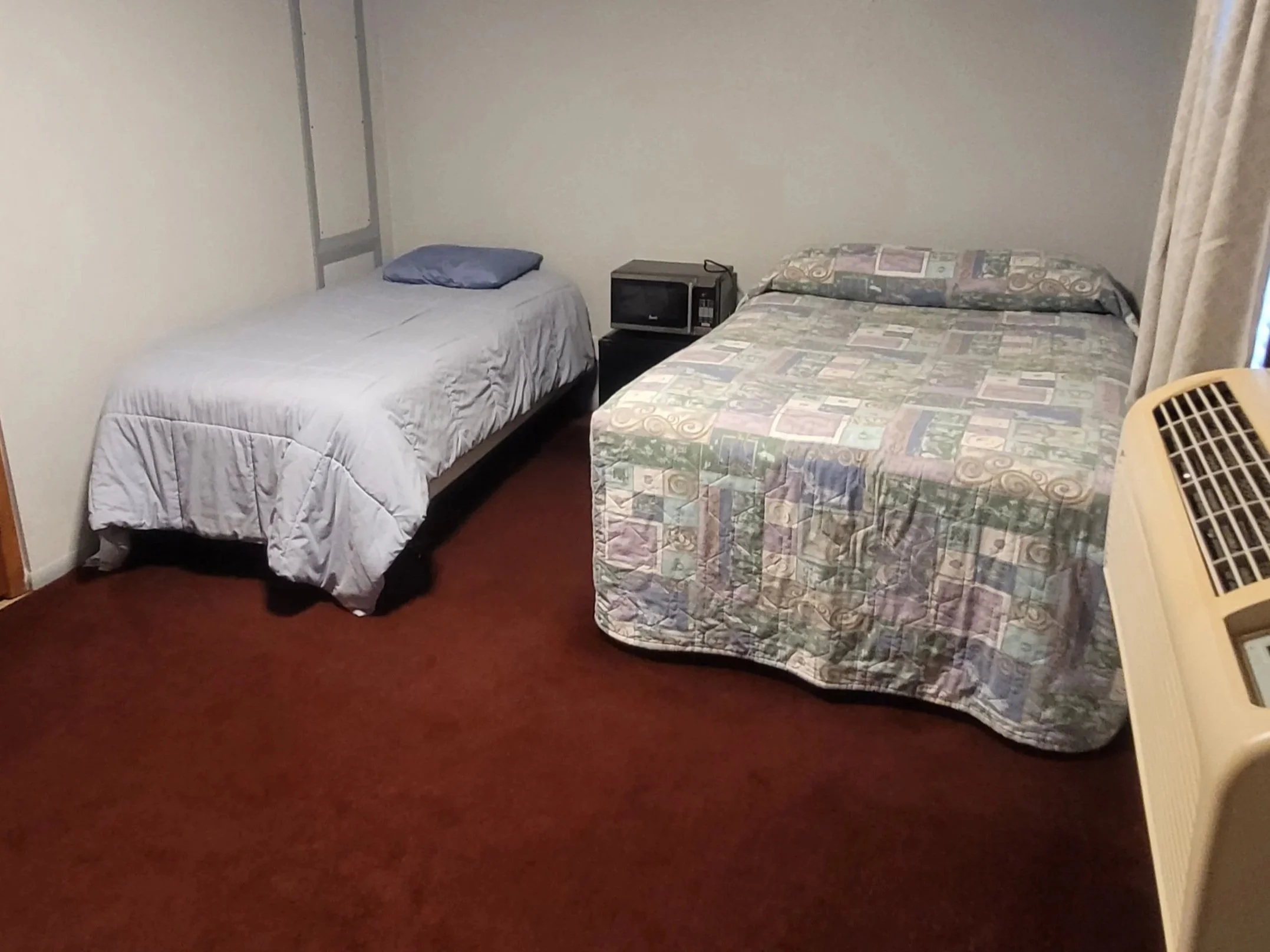 A room with two beds, one with a gray/white quilt and the other with a patterned quilt, a microwave on a small table, a window with curtain, and an air conditioning unit.