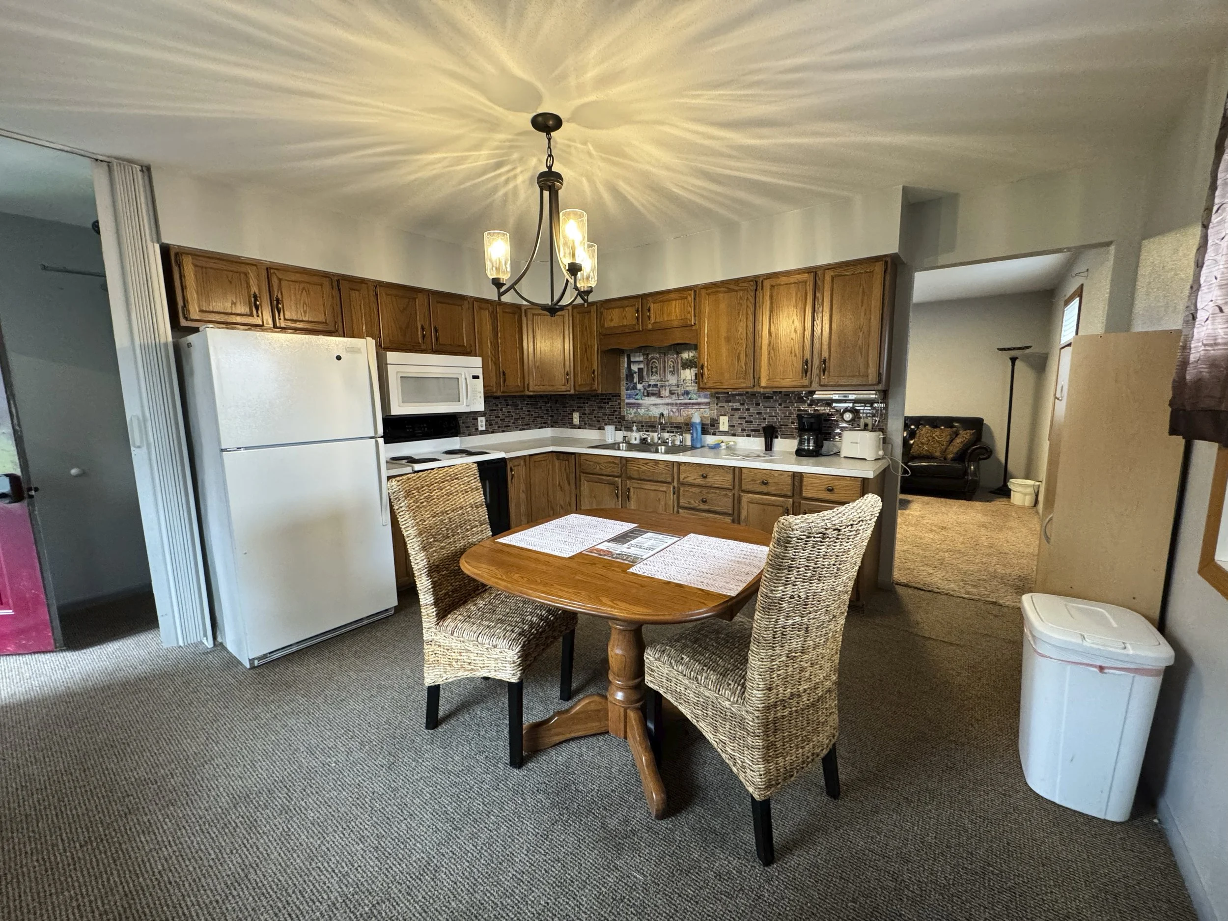 Kitchen with wooden cabinets, white appliances, a round wooden dining table with two woven chairs, a chandelier, and a view into a living room.
