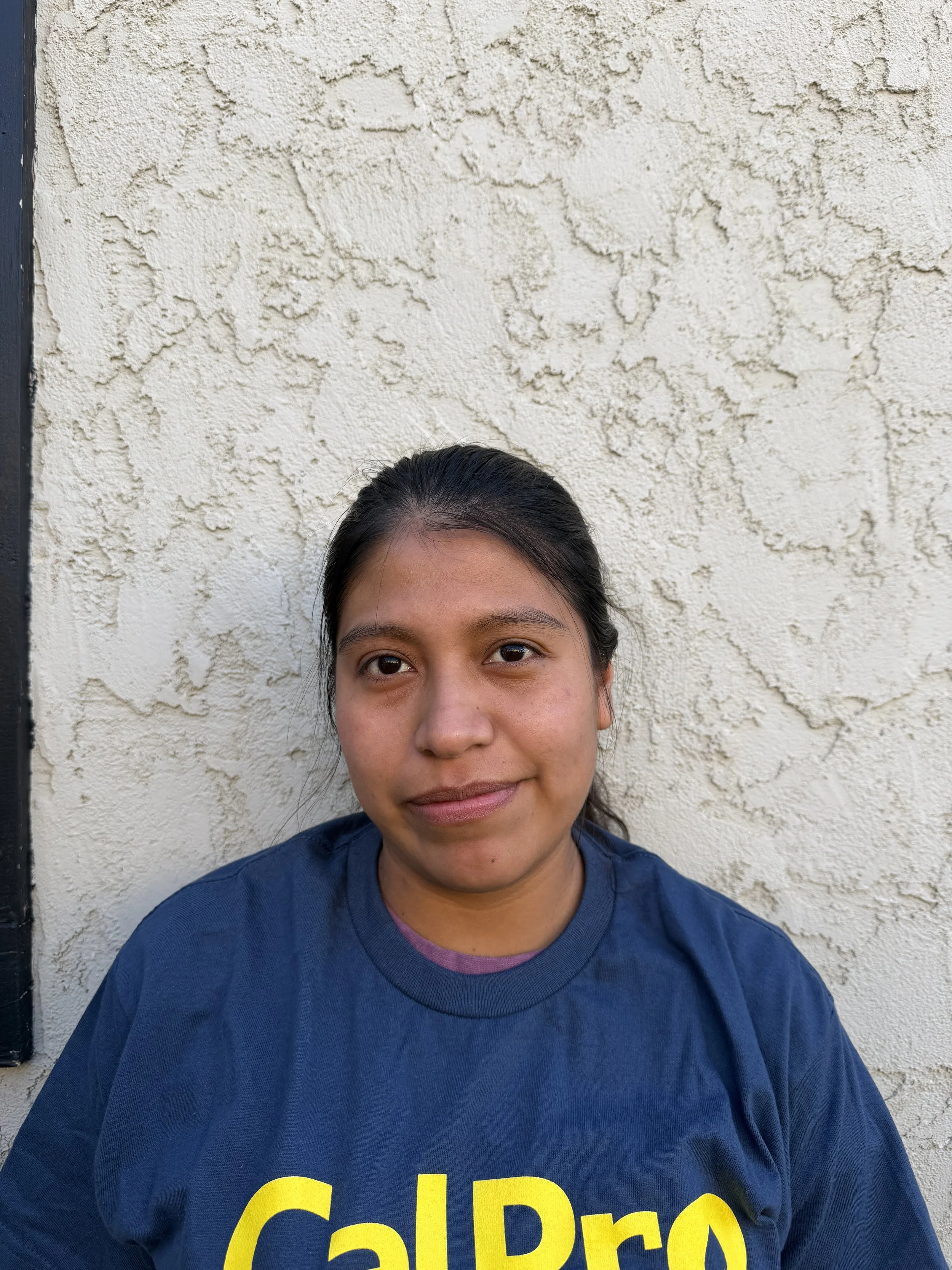 Portrait of a young man standing against a plain off-white wall, wearing a navy blue t-shirt with yellow and white text that reads 'CalPro property care'.