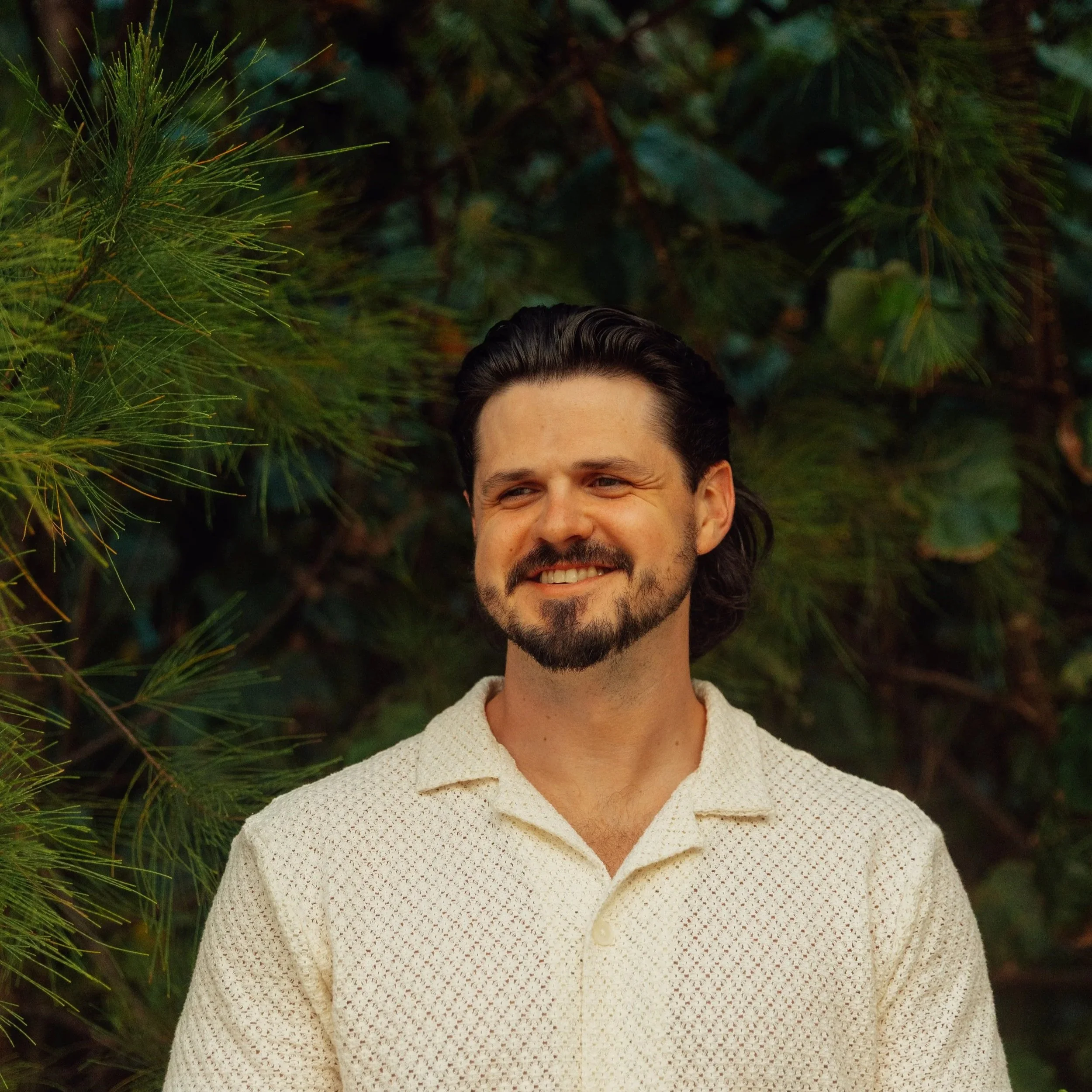 A smiling man with dark, wavy hair and a beard, wearing a light-colored collared shirt, standing outdoors in front of green foliage.