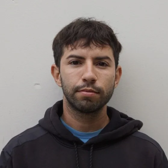 A young man with dark hair and light skin wearing a black hoodie against a plain light gray wall.