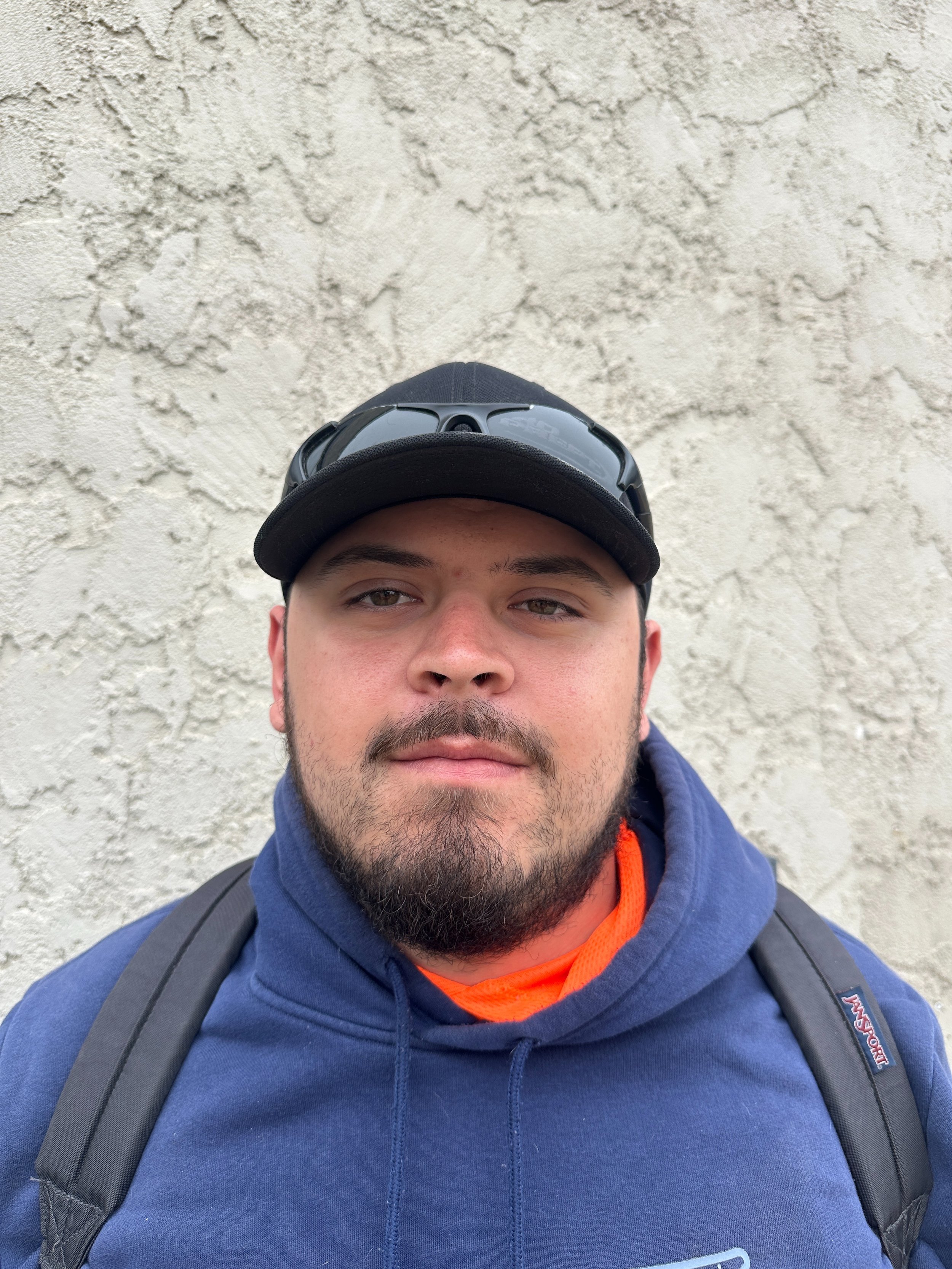 A young man with a beard and mustache wearing a black cap, sunglasses on top of the cap, a blue hoodie, and carrying a backpack, standing in front of a textured white wall.