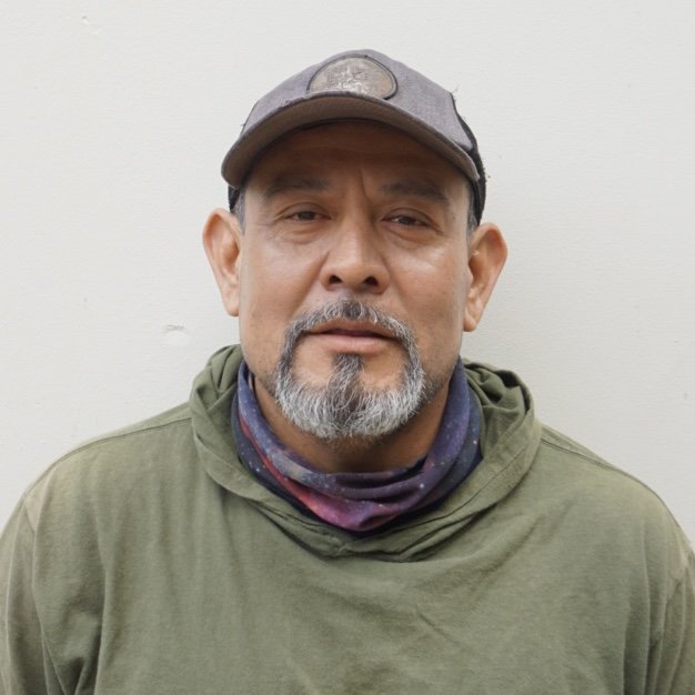 A middle-aged man with a beard and mustache wearing a gray cap, green hoodie, and a colorful neck gaiter, standing against a plain white wall.