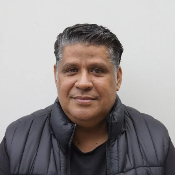 A man with dark hair and tan skin wearing a black puffer jacket and black shirt, smiling slightly against a plain white wall.