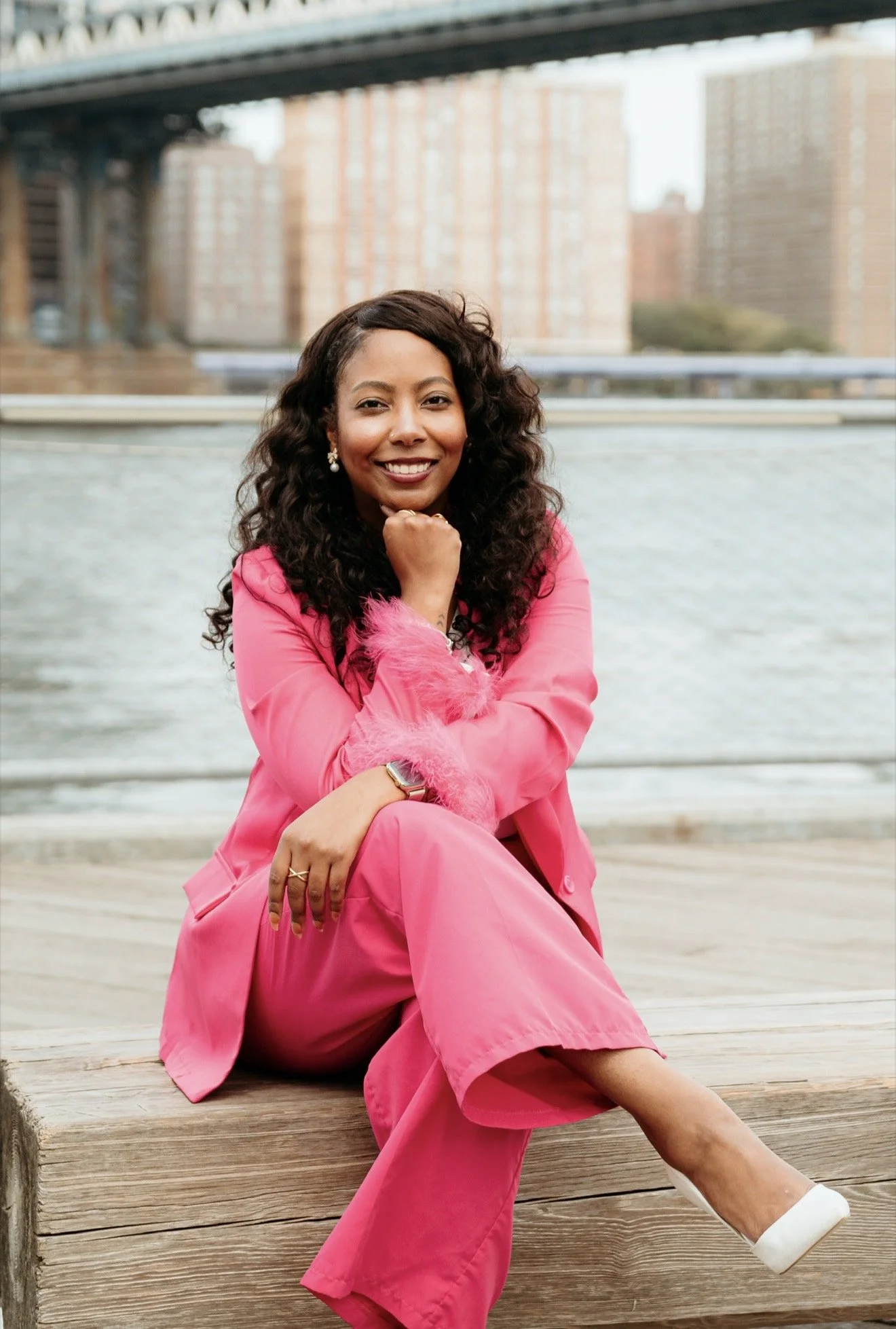A woman with dark curly hair smiling, sitting on a wooden dock near water, wearing a bright pink suit and white shoes with a city bridge and buildings in the background.