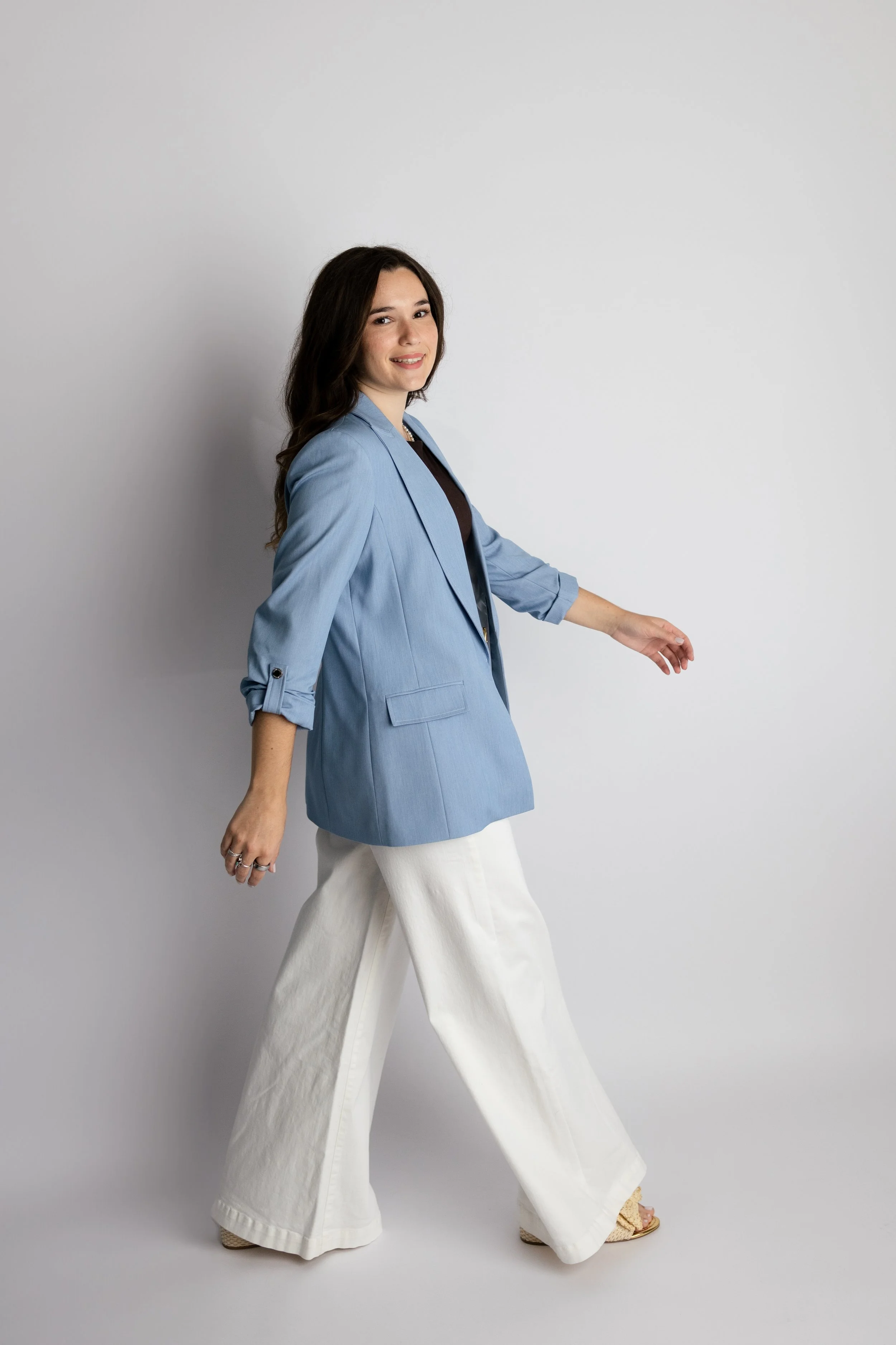 A young woman walking, wearing a light blue blazer, white wide-leg pants, and sandals, against a plain white background.