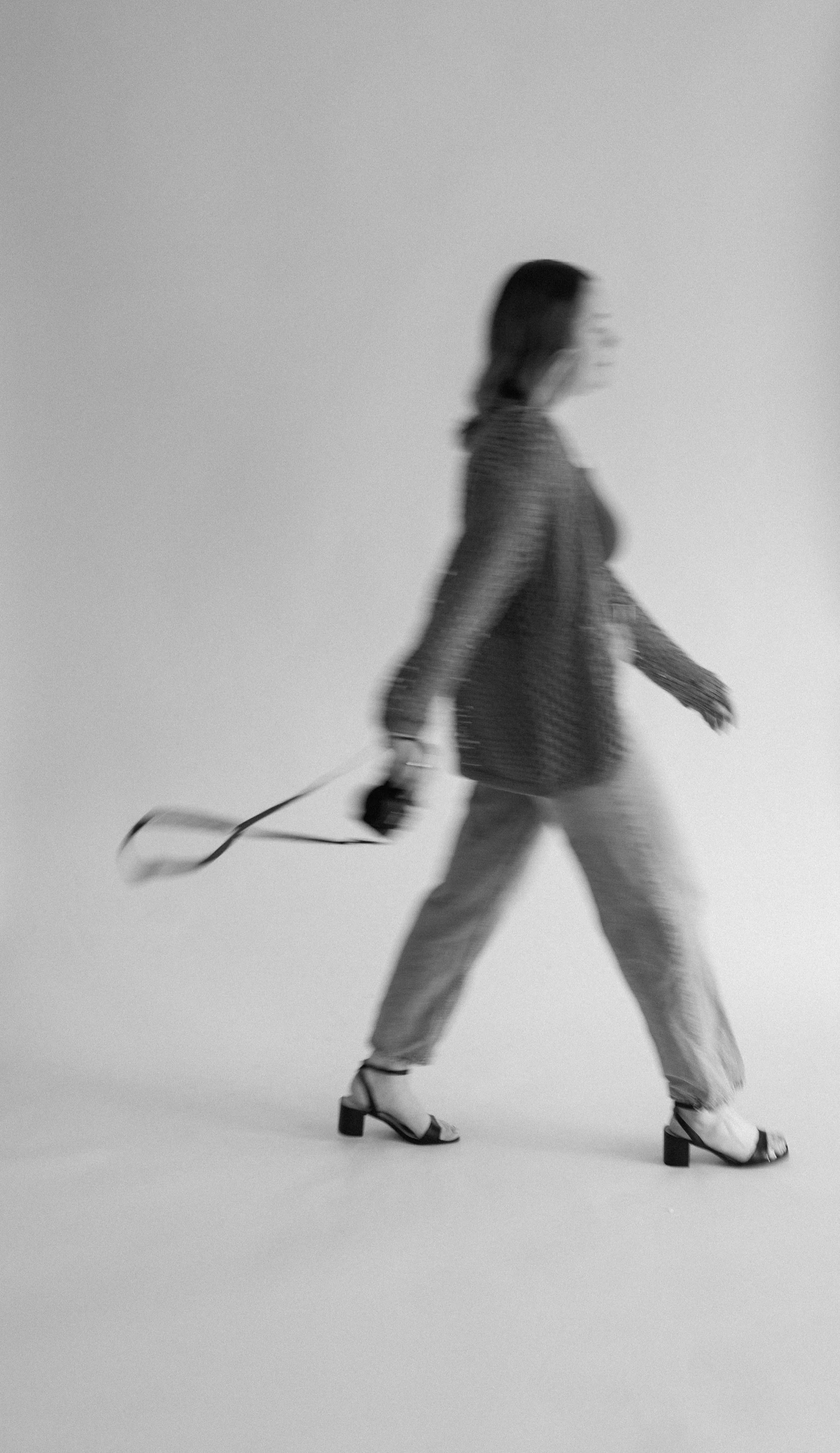 A woman walking with a camera in her right hand, wearing a cardigan, light-colored pants, and black heeled shoes, against a plain background.