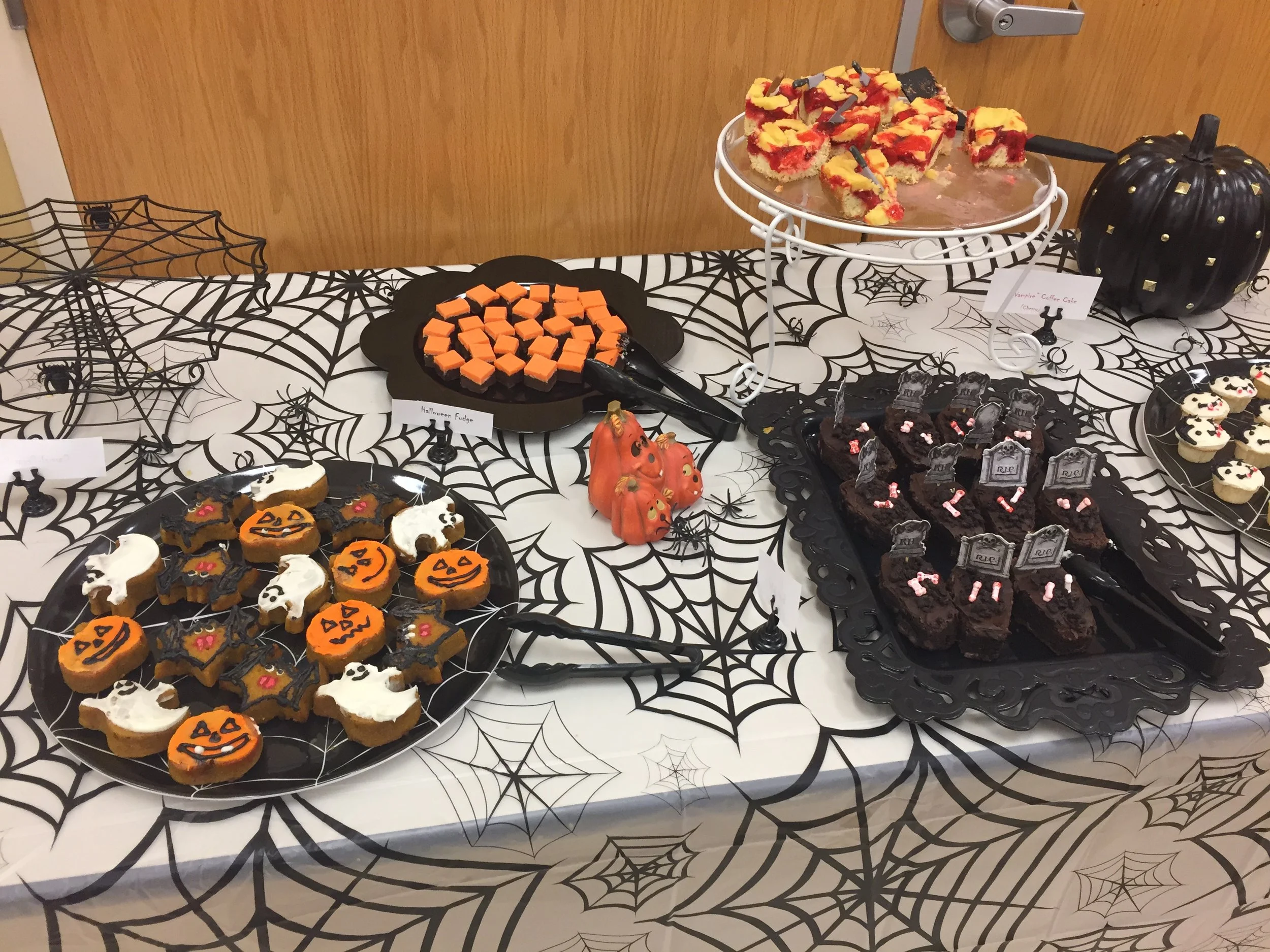 Table decorated with Halloween-themed treats, including cookies with orange jack-o'-lantern faces, ghost-shaped cookies with white icing, and brownies with pink and white candies, set on a spiderweb-patterned tablecloth with Halloween decorations.