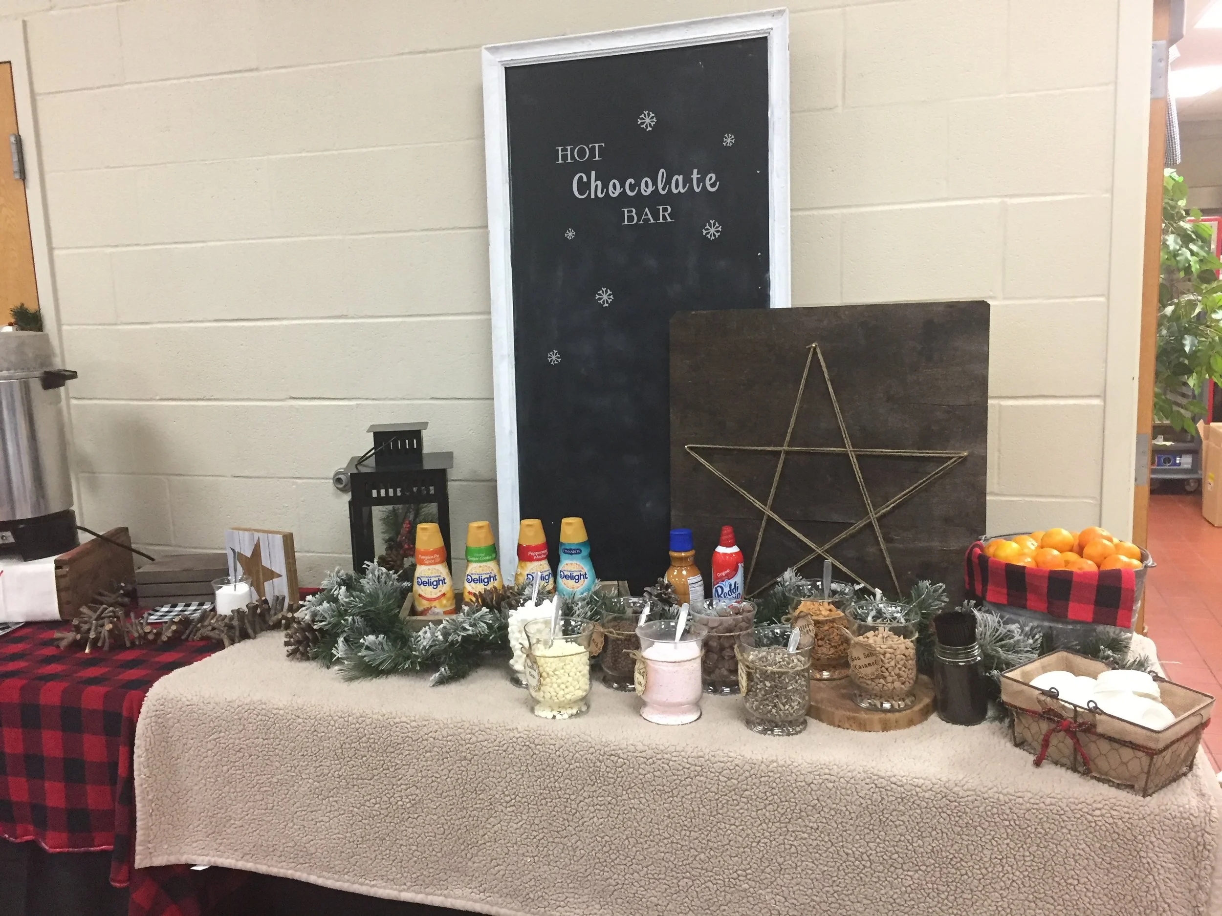 A chocolate bar stand with a black sign reading 'Hot Chocolate Bar' surrounded by small snowflake stickers, and various toppings and condiments on a table decorated with a festive holiday garland, candies, and snacks.