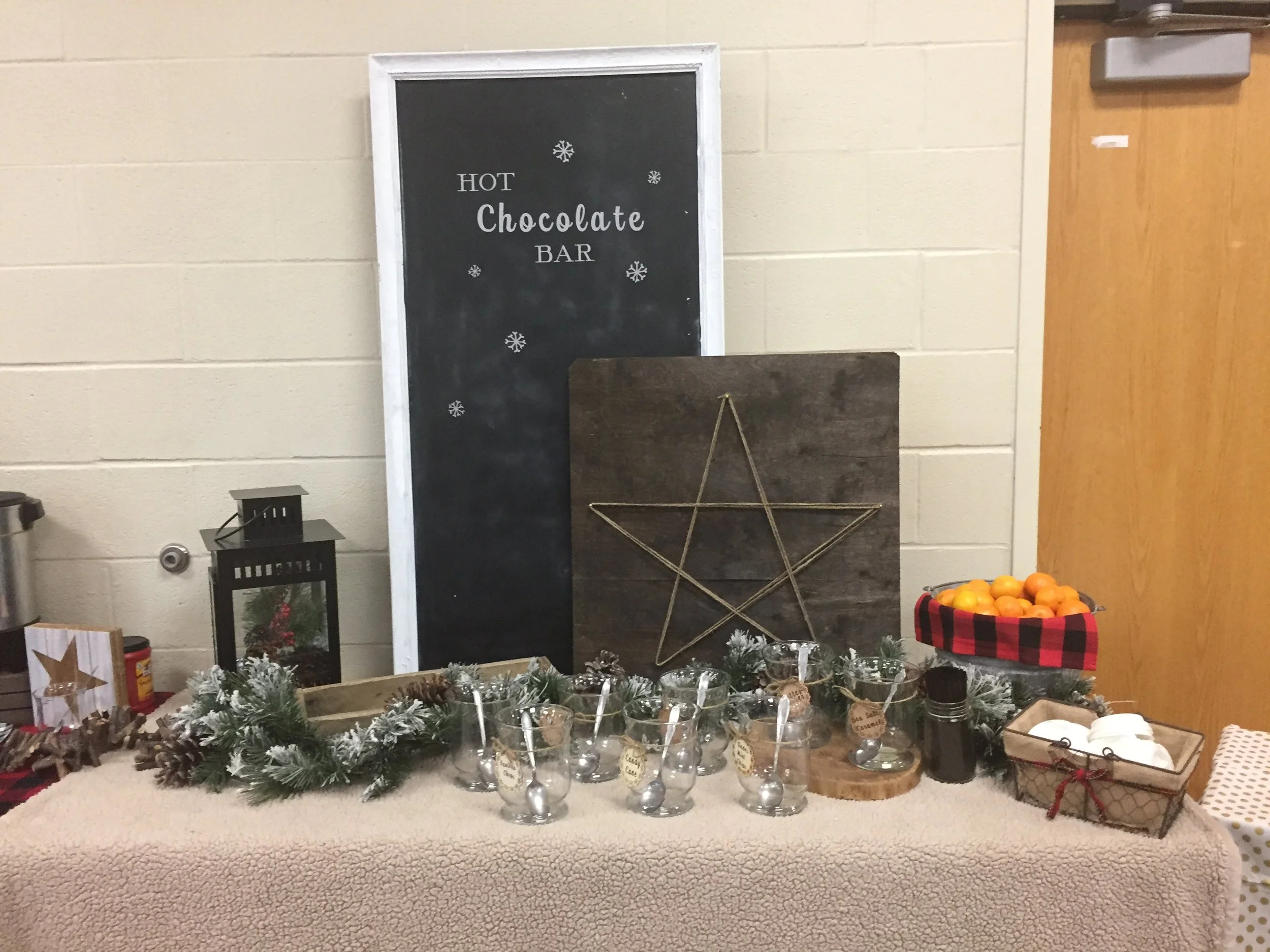 A holiday display with a black chalkboard sign reading 'Hot Chocolate Bar' decorated with snowflakes, and a table with various cups, sprigs of evergreen, pinecones, and festive decor, alongside a bowl of oranges, set against a beige wall and a wooden