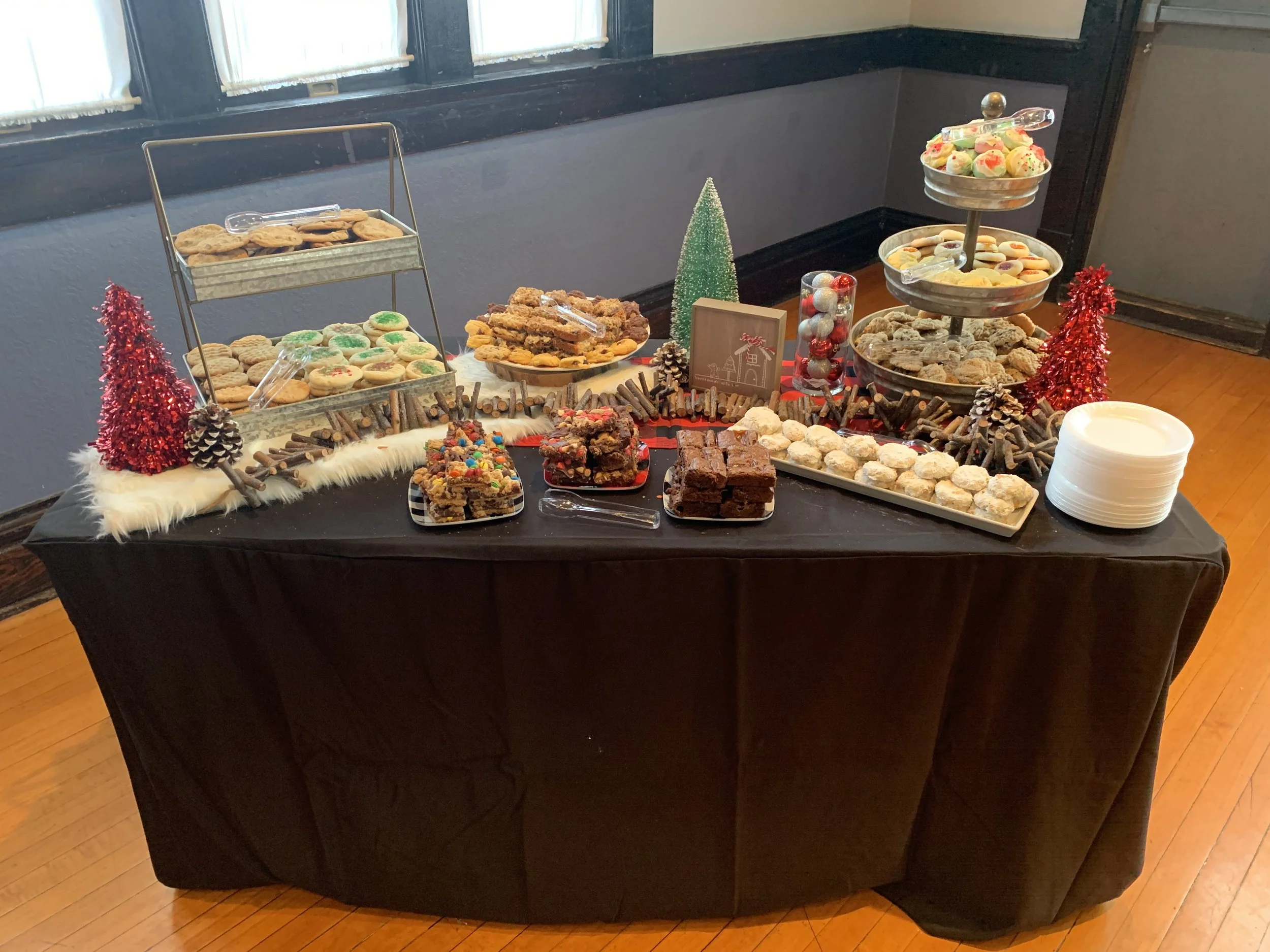 A dessert table with cookies, brownies, and candies, decorated with small red and green Christmas trees, pinecones, and holiday-themed decor.
