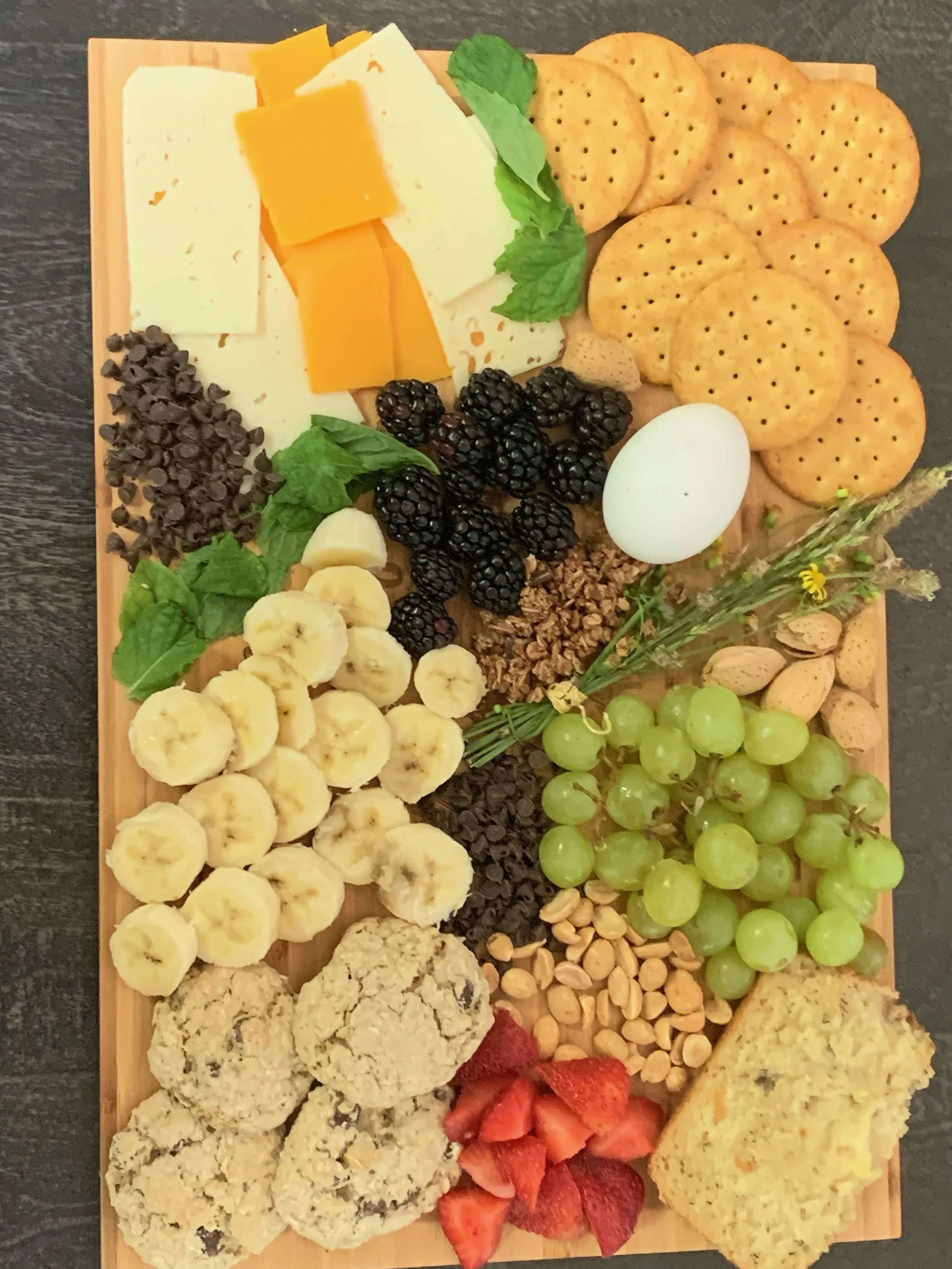 A cheese and fruit platter with various cheeses, grapes, strawberries, strawberries, sliced bananas, blackberries, nuts, and crackers arranged on a wooden board.