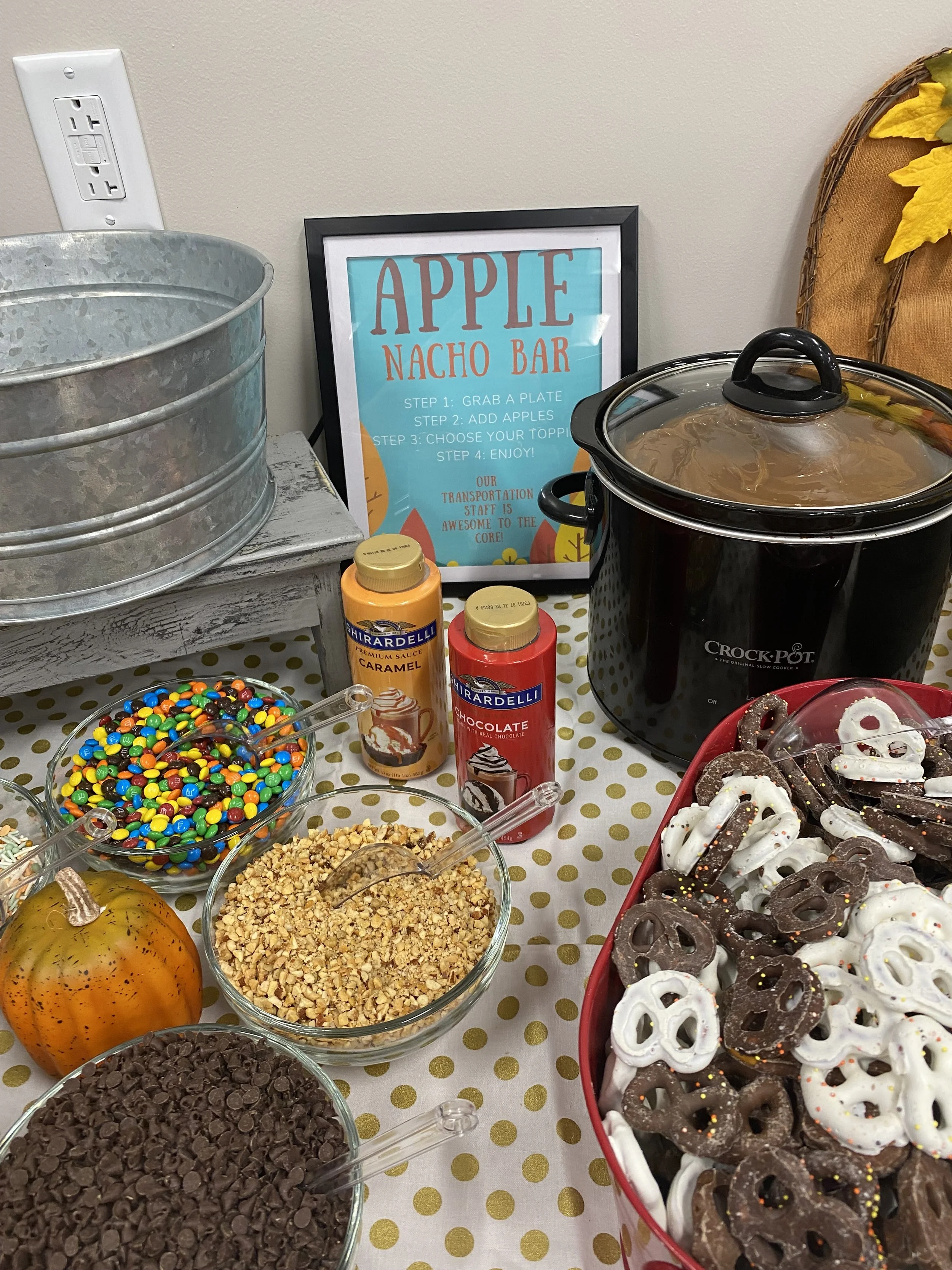A dessert table featuring a black crockpot, bowls of various toppings and candies including chocolate chips, M&Ms, crushed nuts, and sprinkles, a pumpkin, and a sign that reads 'Apple Nacho Bar' with instructions for making apple nachos, alongside a 