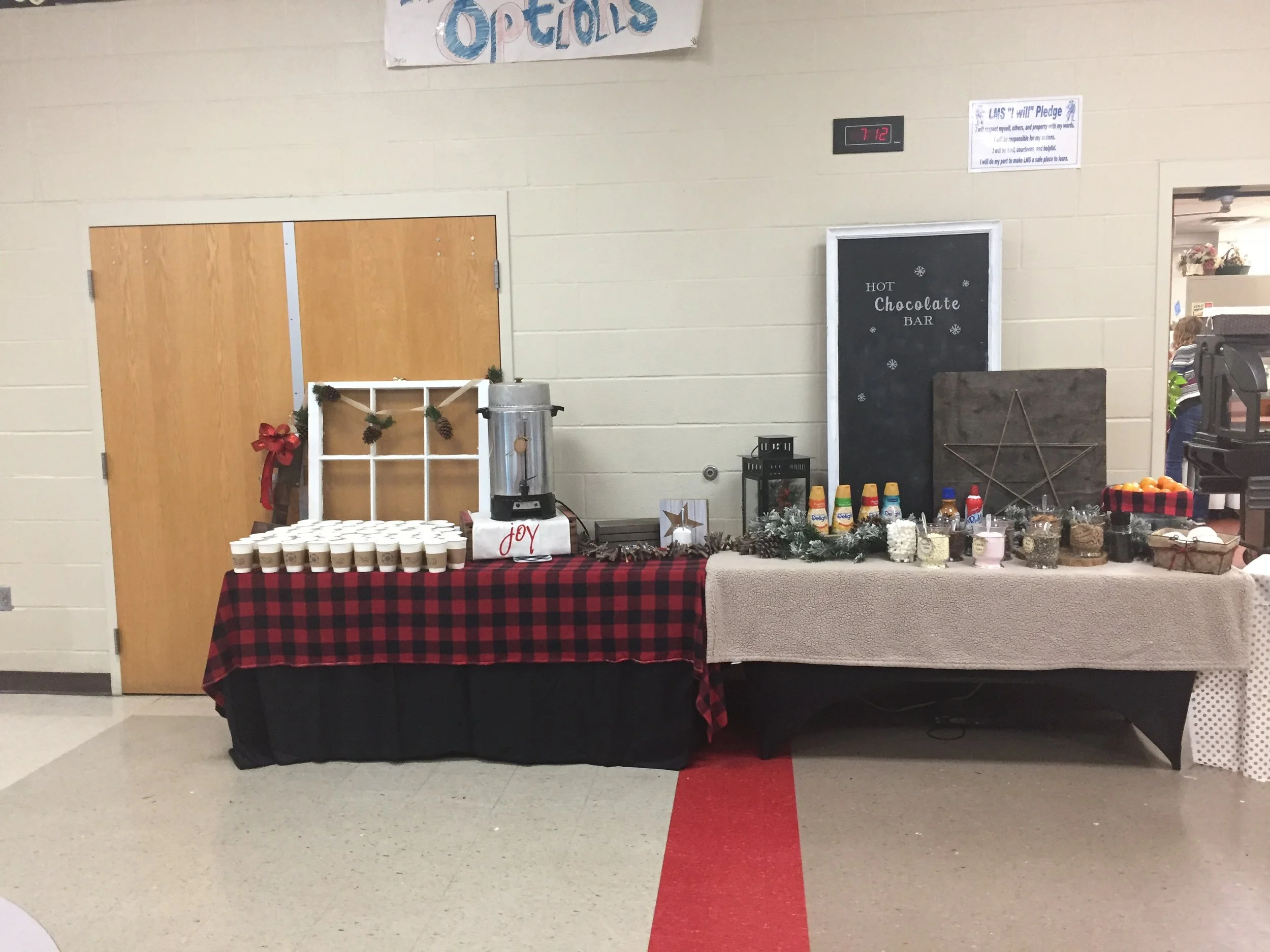 A holiday-themed beverage station with cups, a coffee dispenser, and assorted supplies on a table covered with a red and black checkered tablecloth. A sign reads “HOT Chocolate BAR,” and there are Christmas decorations including pinecones, ornaments,