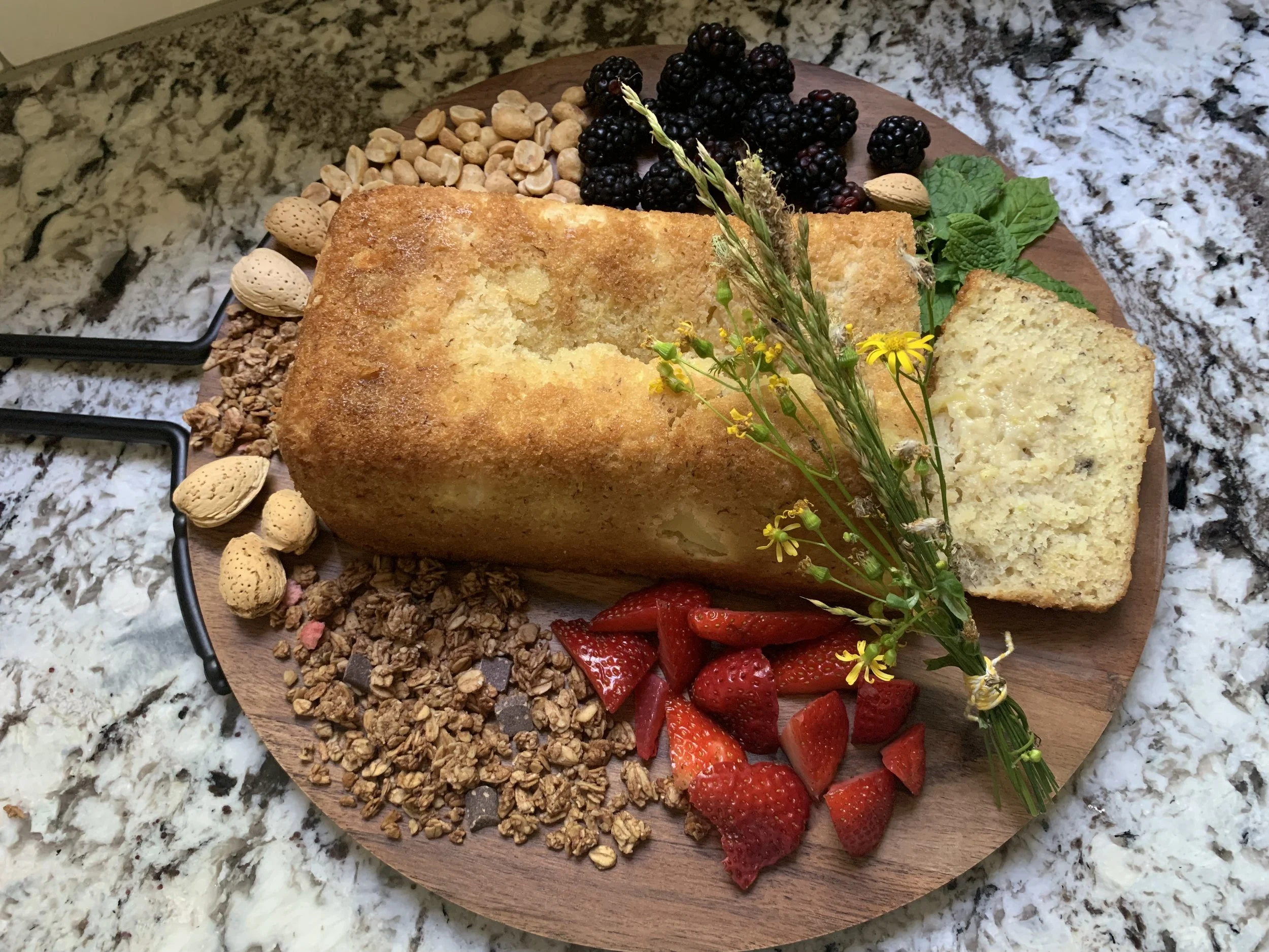 A wooden serving board with a sliced pound cake, fresh strawberries, blackberries, almonds, peanuts, granola, chocolate chunks, and a sprig of herbs and yellow flowers on top, placed on a granite countertop.