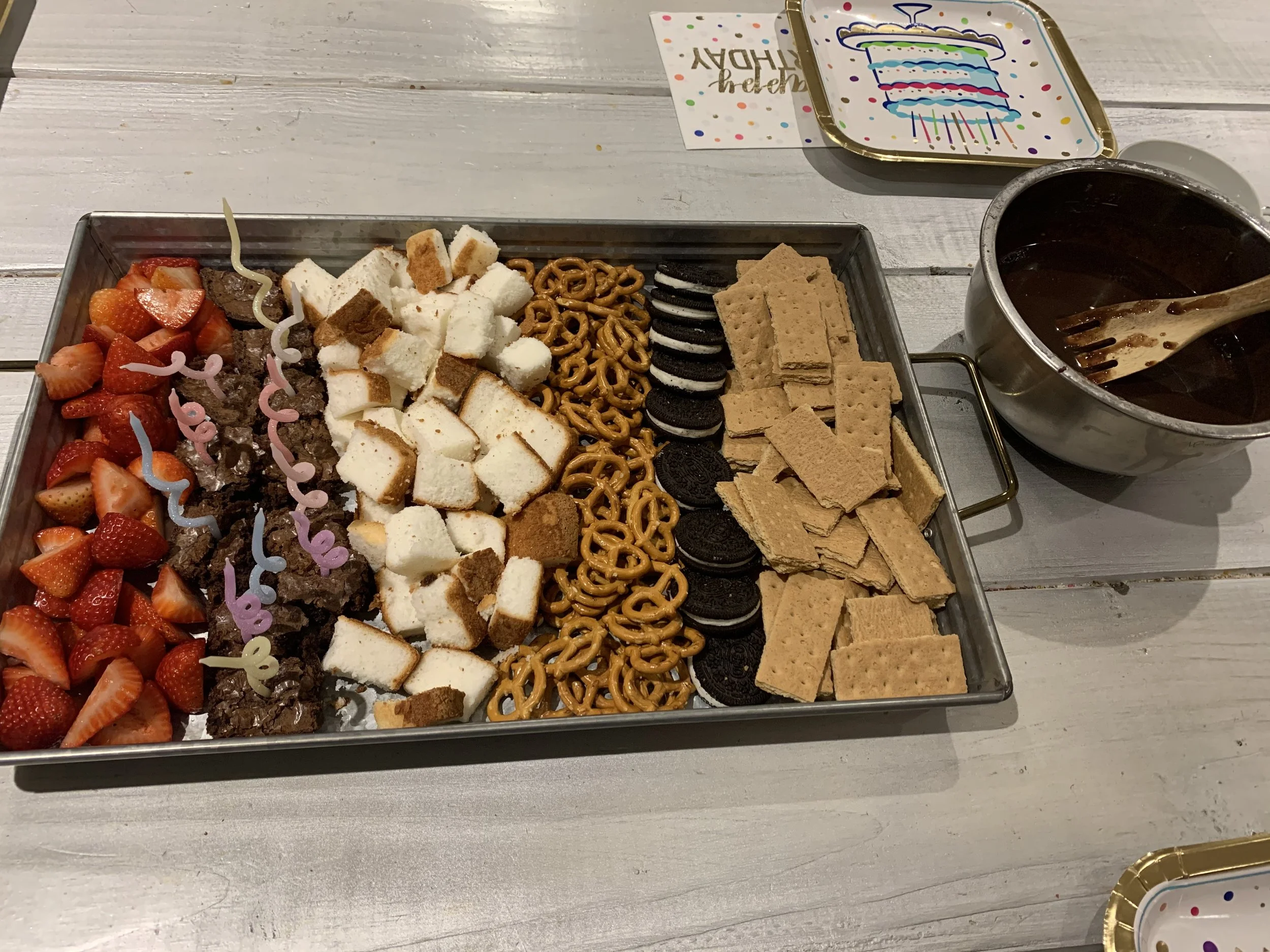 A celebration platter with fresh strawberries, chocolate candies, cake cubes, pretzels, Oreos, graham crackers, and a cup of dark beverage with a spoon, on a white wooden table next to party plates and a "happy birthday" card.