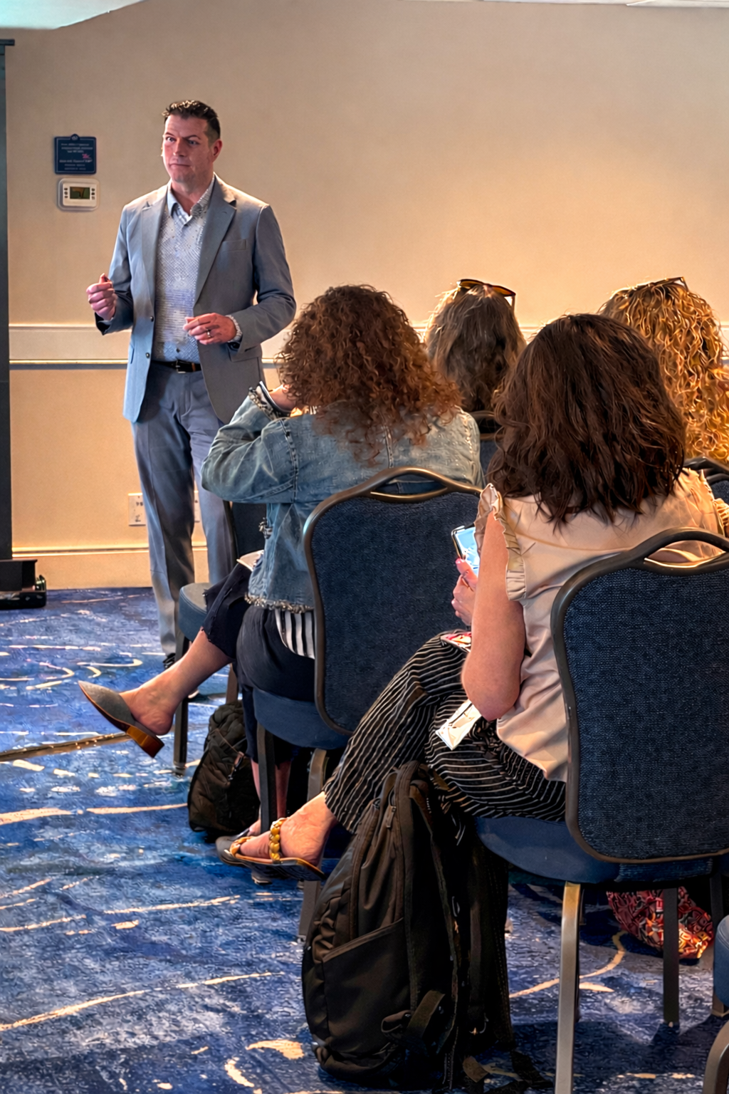 A man in a grey suit standing and speaking to a small audience of women seated in chairs, some using their phones, in a conference room with a blue carpet.