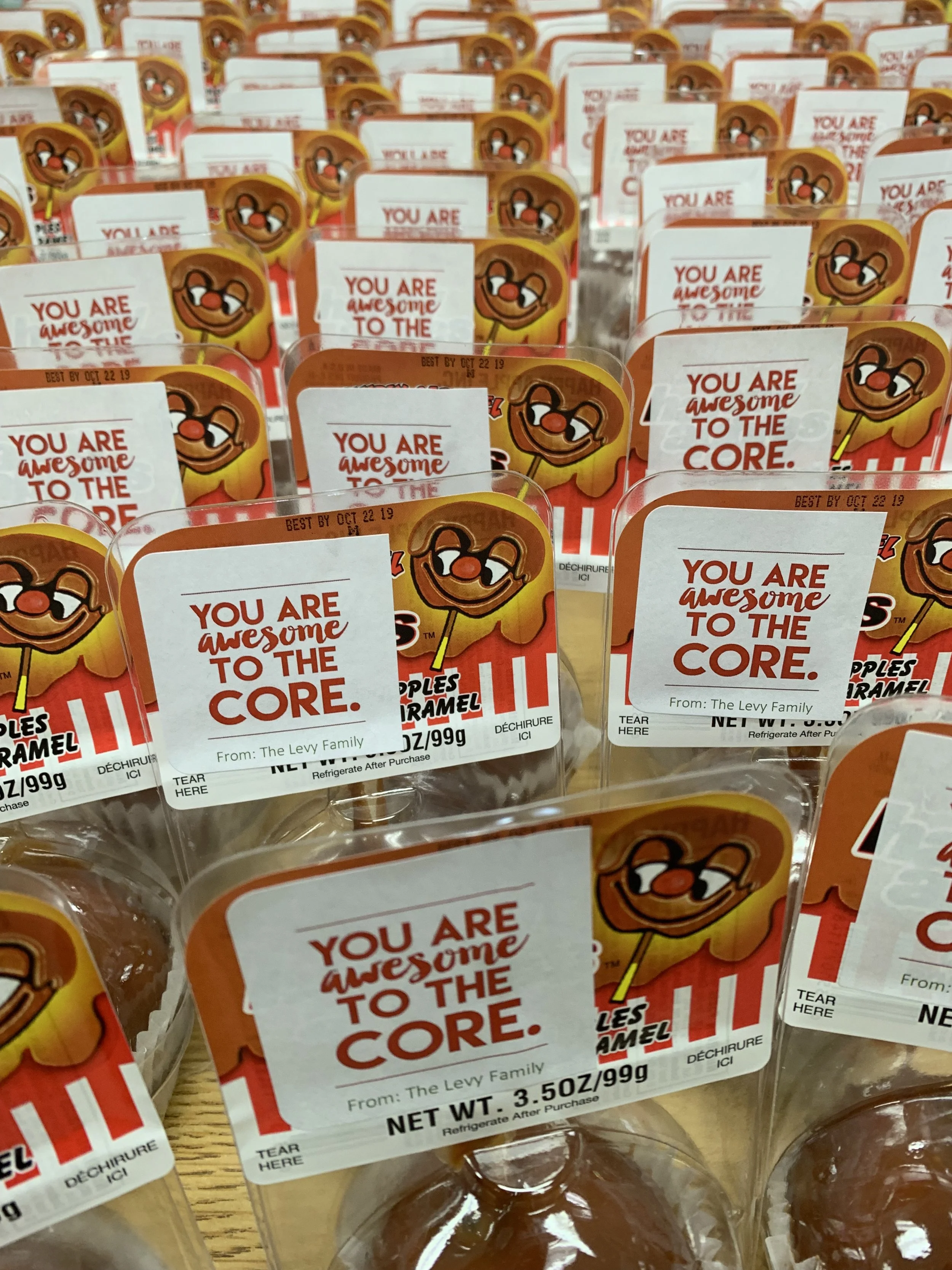 Multiple packages of caramel apples with a label that reads 'You are awesome to the core,' featuring a cartoon smiling caramel apple character with a red nose and a stick. The packages are arranged on a display shelf.