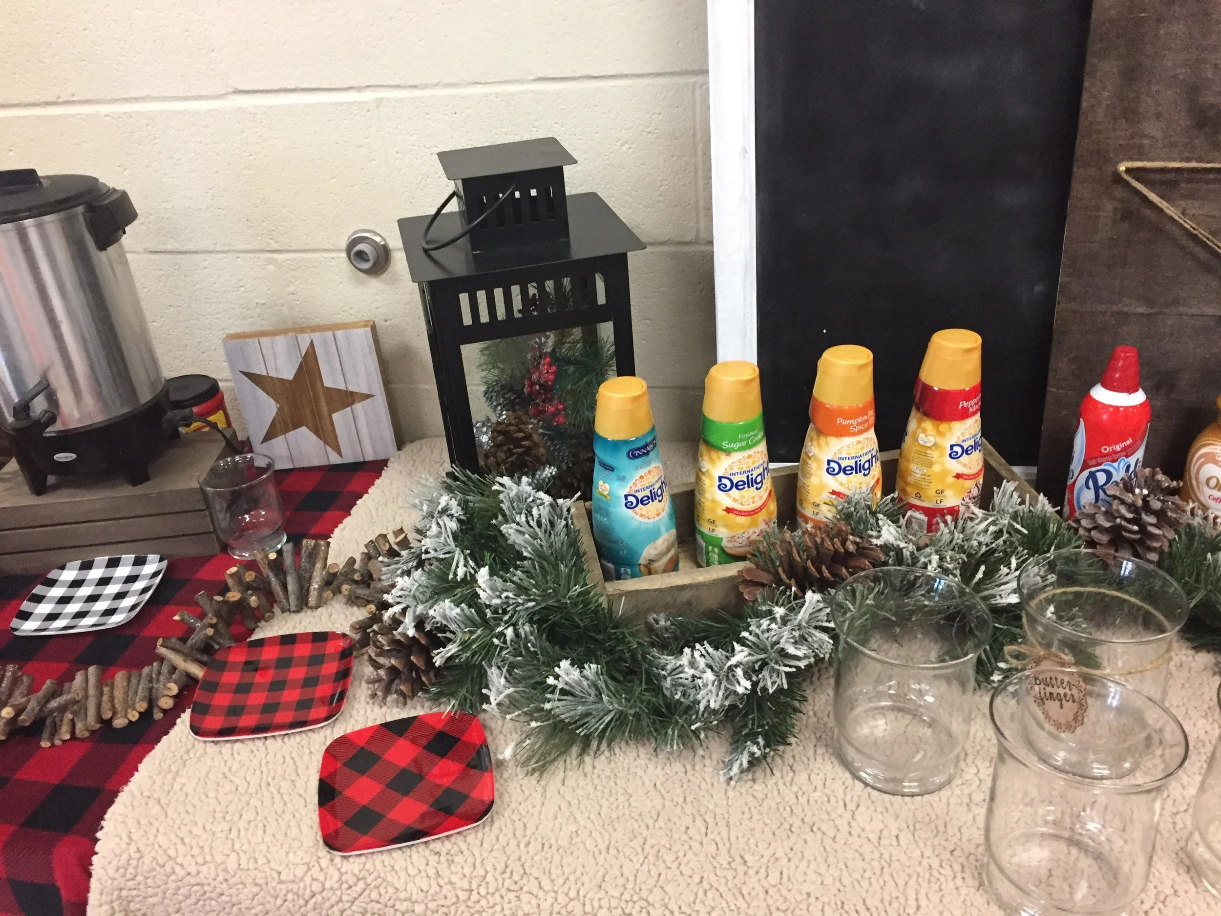 Table decorated with Christmas-themed items, including plaid napkins, a small artificial Christmas tree, candles, and pinecones, with a black lantern and a wall in the background.