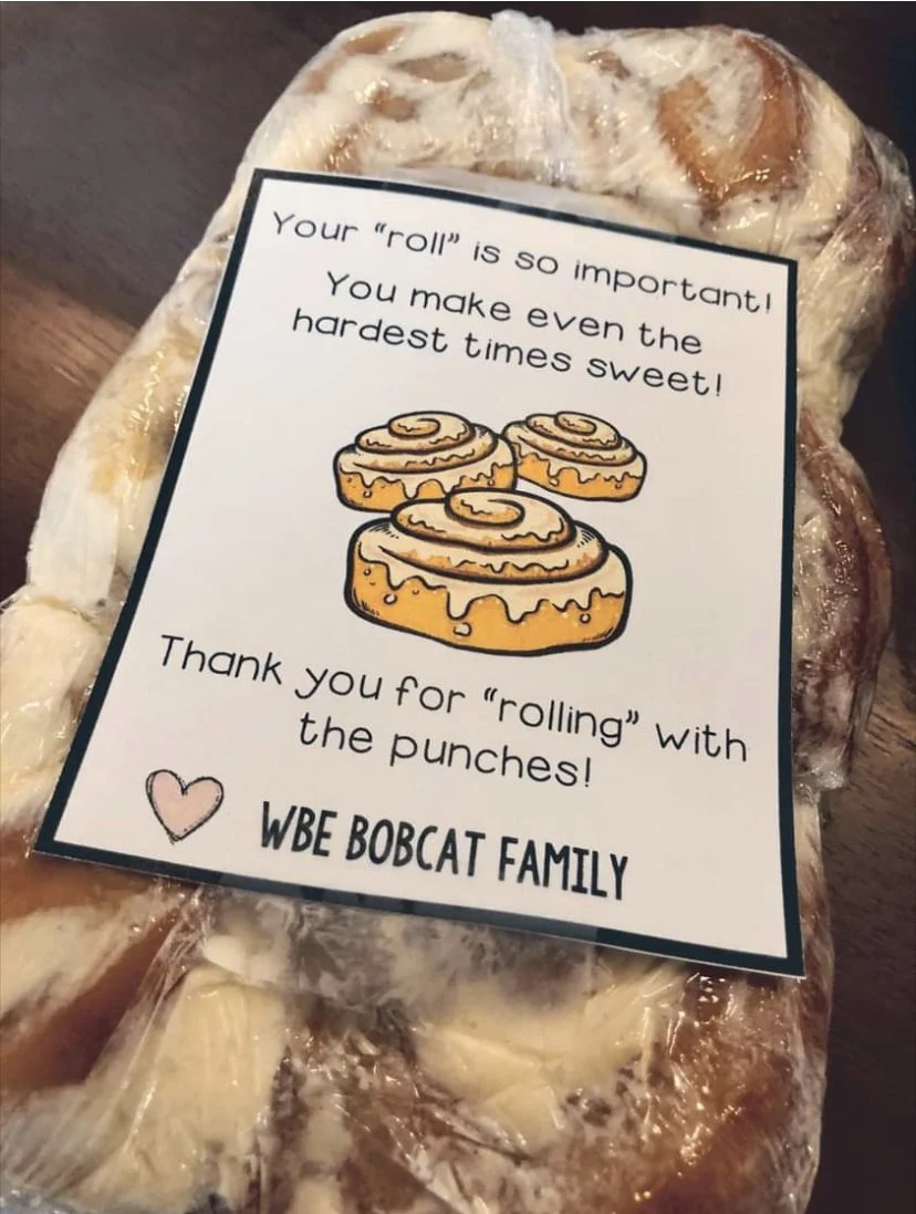 A cinnamon roll cake wrapped in clear plastic with a note attached to it. The note has a message about the importance of cinnamon rolls and includes a drawing of three cinnamon rolls. It is signed by the WBE Bobcat Family.