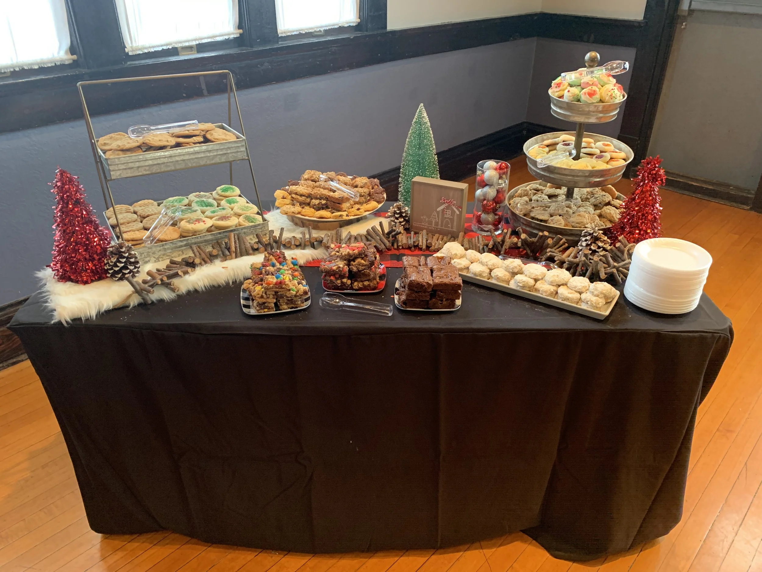 A dessert table featuring cookies, brownies, and assorted sweets arranged on tiered trays and plates, decorated with small red and green Christmas trees, pinecones, and holiday-themed accents.