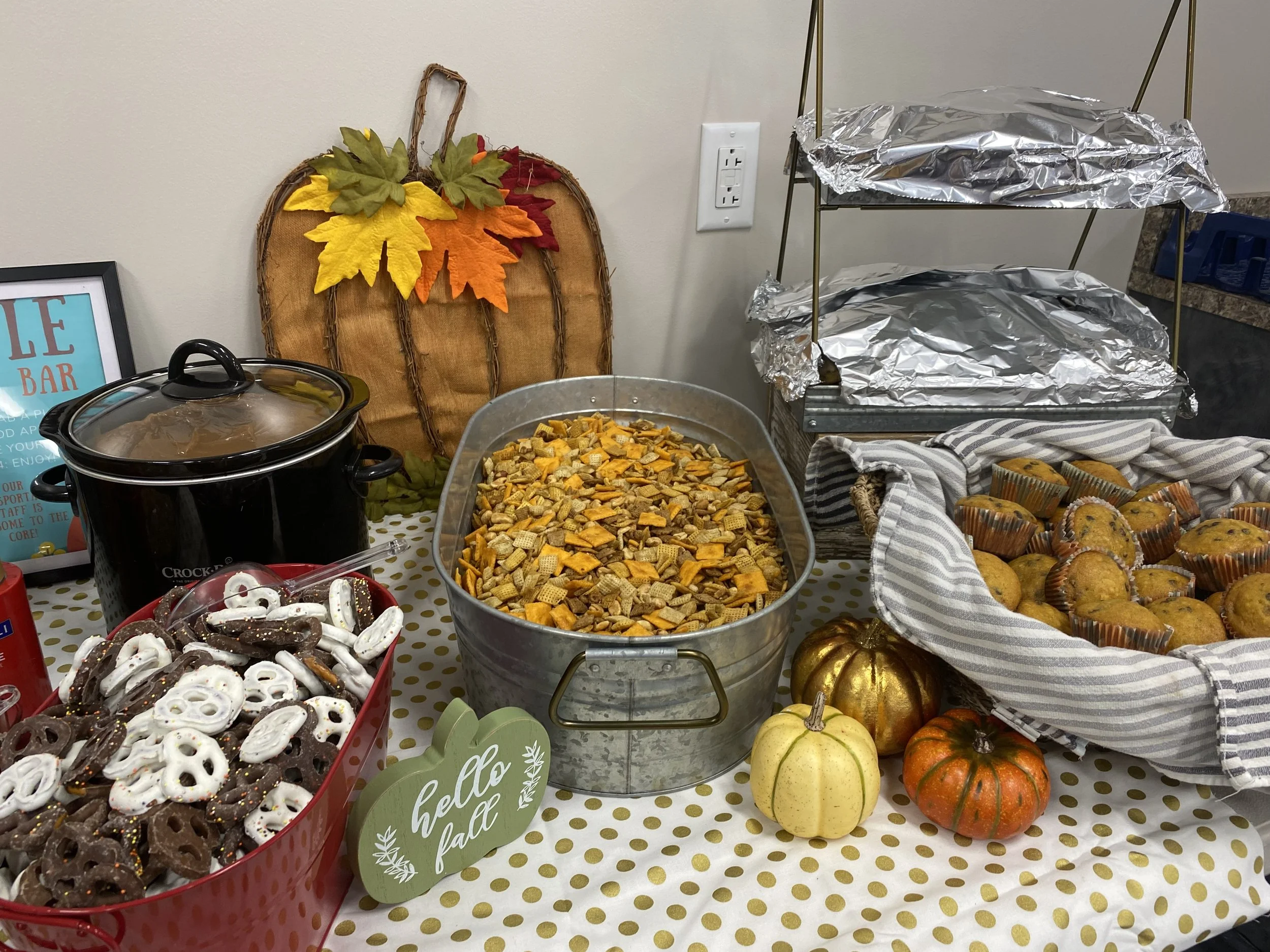 A table with a variety of fall-themed treats and decorations including a tray of cereal snack mix, chocolate-covered pretzels, cupcakes, mini pumpkins, and a decorative pumpkin made from painted gourds, with an autumn leaf backdrop and a sign that sa