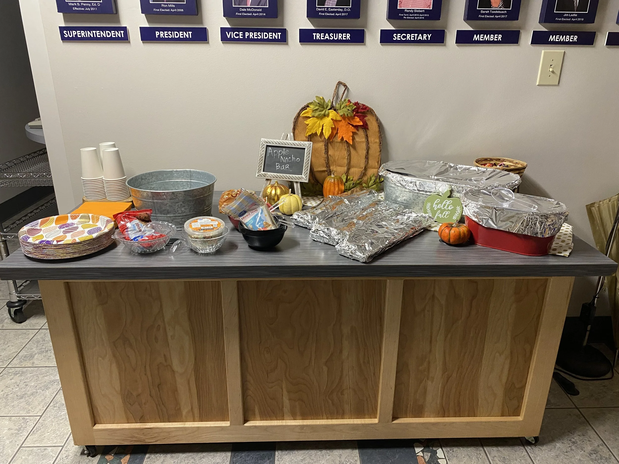 A wooden table with foil-covered dishes, a small chalkboard sign reading 'Apple Nacho Bar', decorative autumn leaves, small pumpkins, disposable cups, a metal bucket, and various bowls of food in a room with wall plaques and a tiled floor.