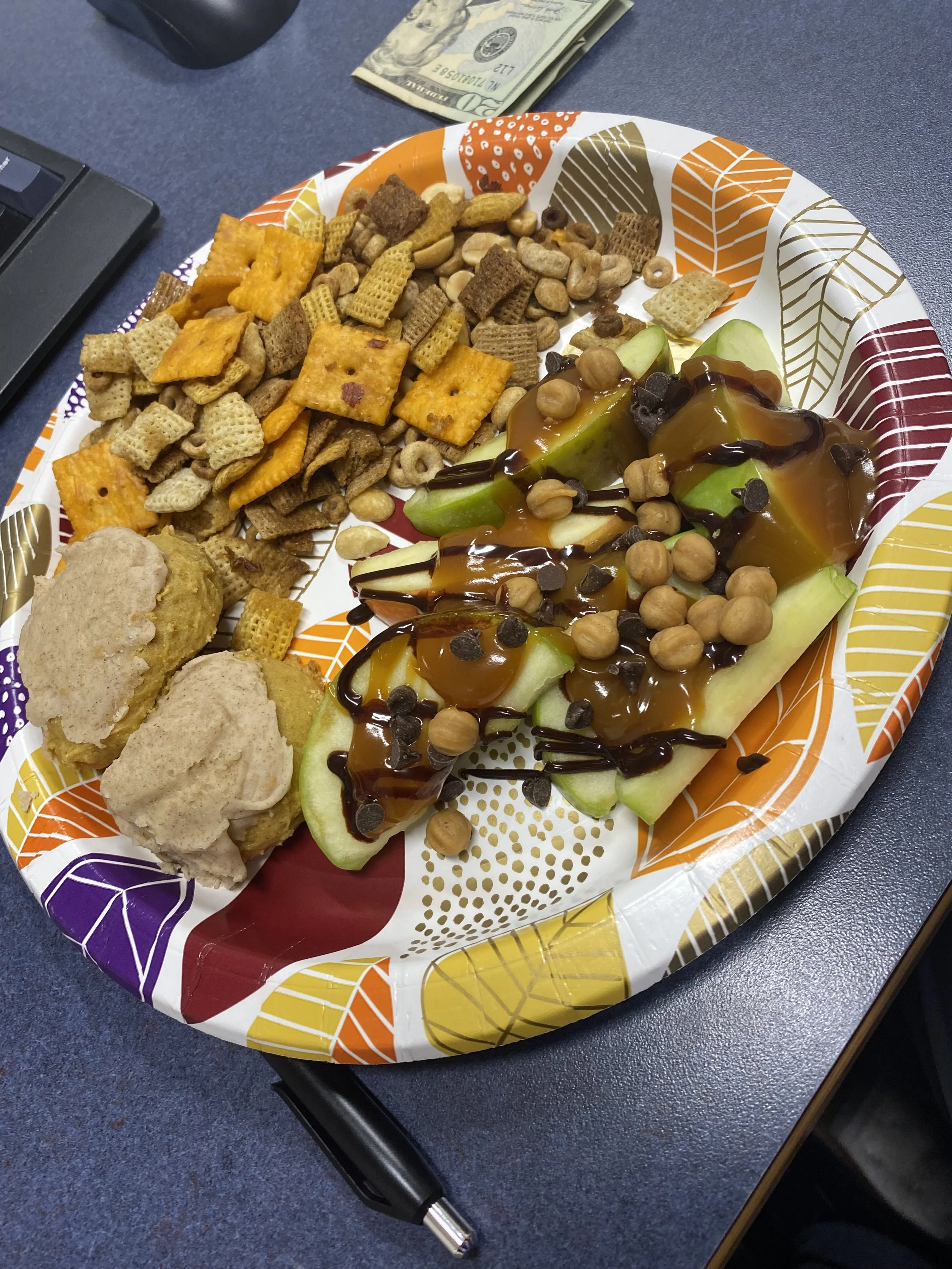 A paper plate with a colorful leaf pattern containing apple slices topped with caramel, chocolate chips, and peanuts, along with trail mix crackers, cereal pieces, and two cookies. A pen and a $20 bill are on a blue table.