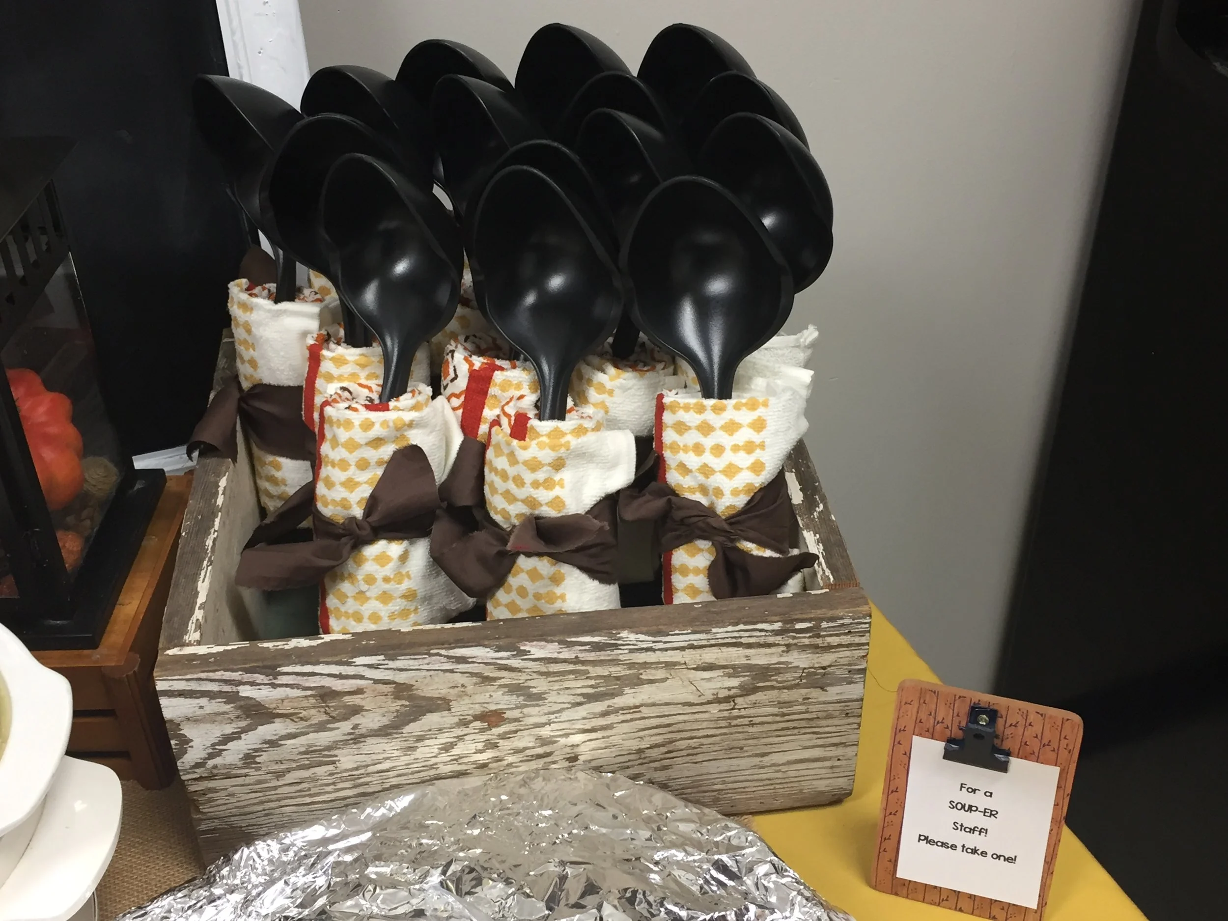 Black plastic soup spoons in paper napkin holders on a rustic wooden table, with a sign requesting diners to take one.