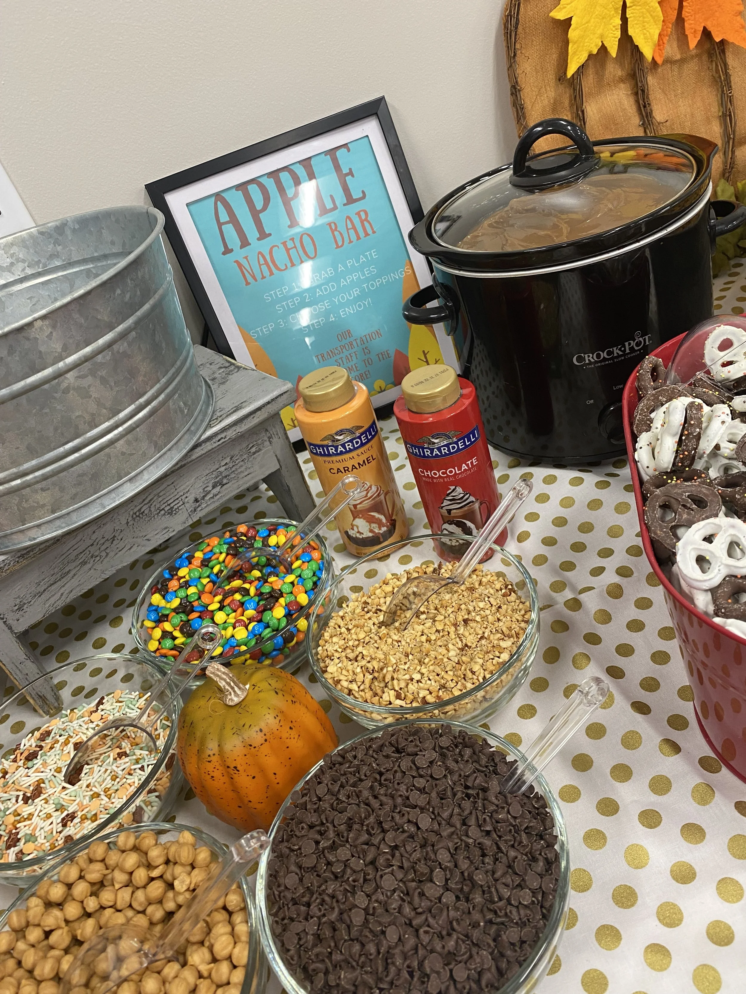 A table with various toppings, including chocolate chips, rainbow sprinkles, chopped nuts, and a small pumpkin, at a dessert or snack station. There are also bottles of caramel and chocolate syrup, a crock-pot, and a sign with instructions for an app