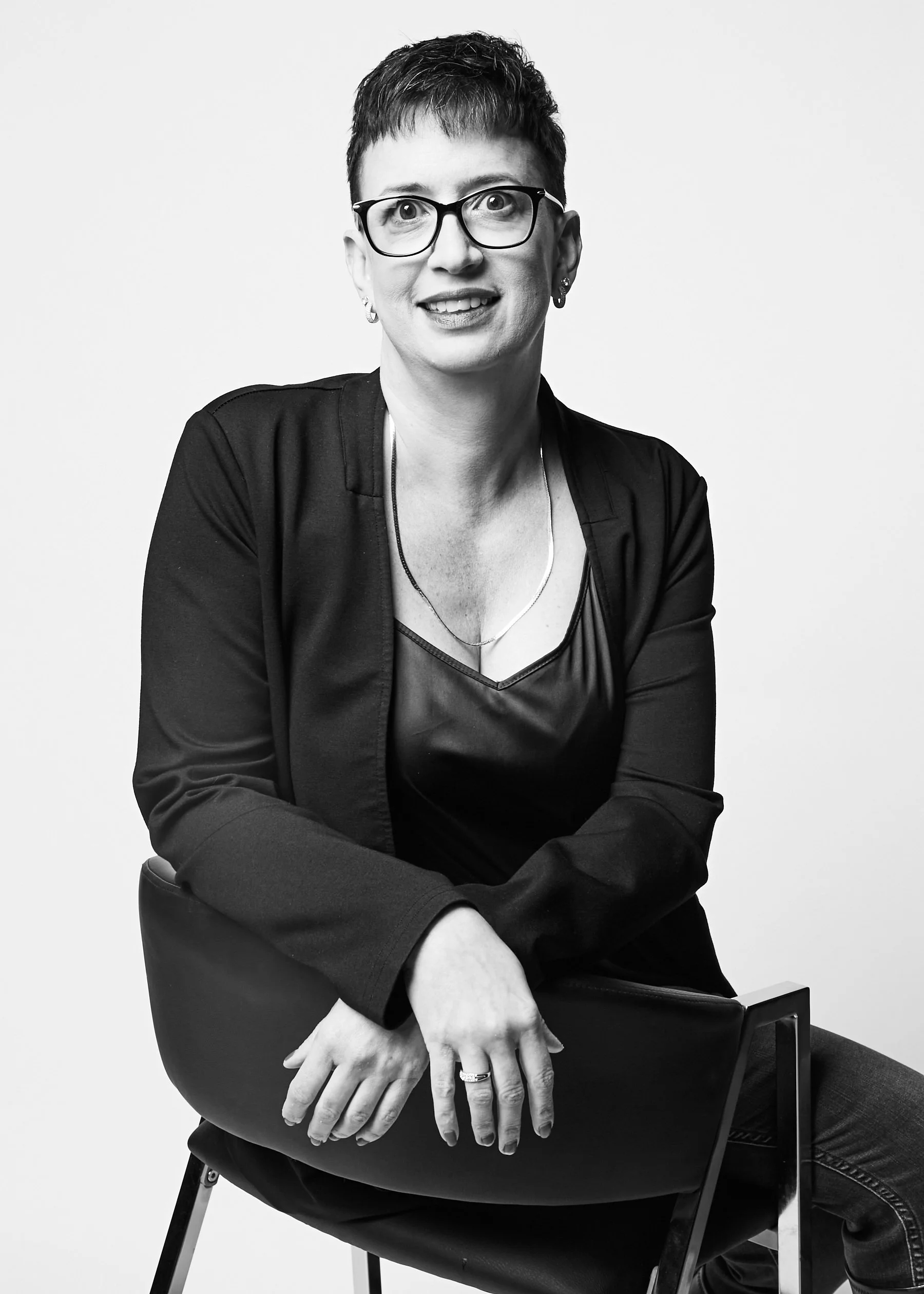 Black and white portrait of a woman with short hair, glasses, and earrings, sitting on a chair, wearing a dark jacket and shirt, looking at the camera with a smile.
