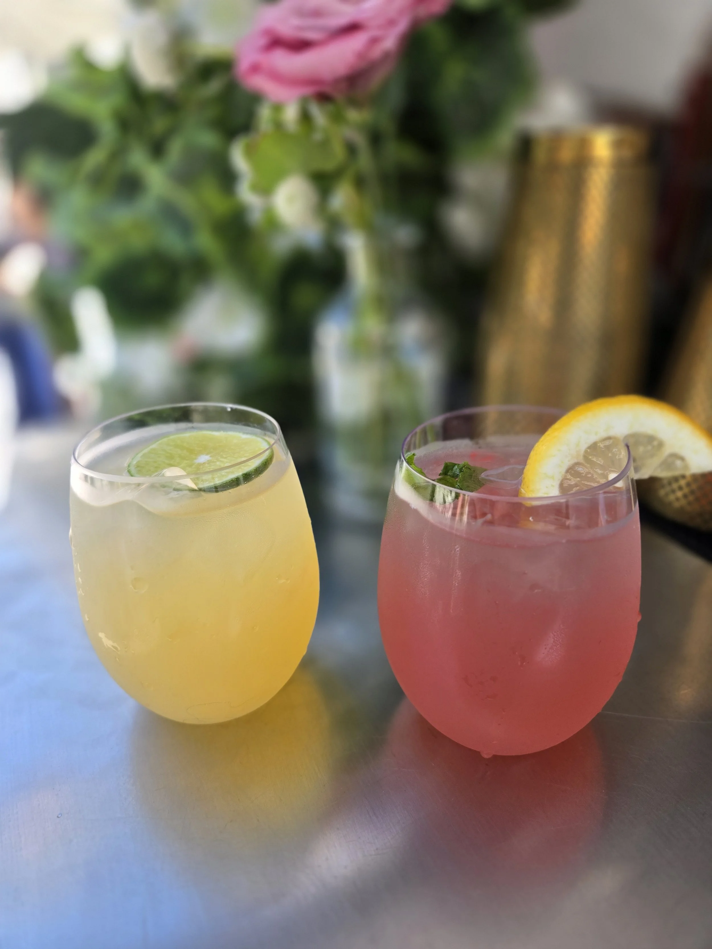 Two colorful cocktails garnished with lime and lemon beside flowers and gold bar tools.