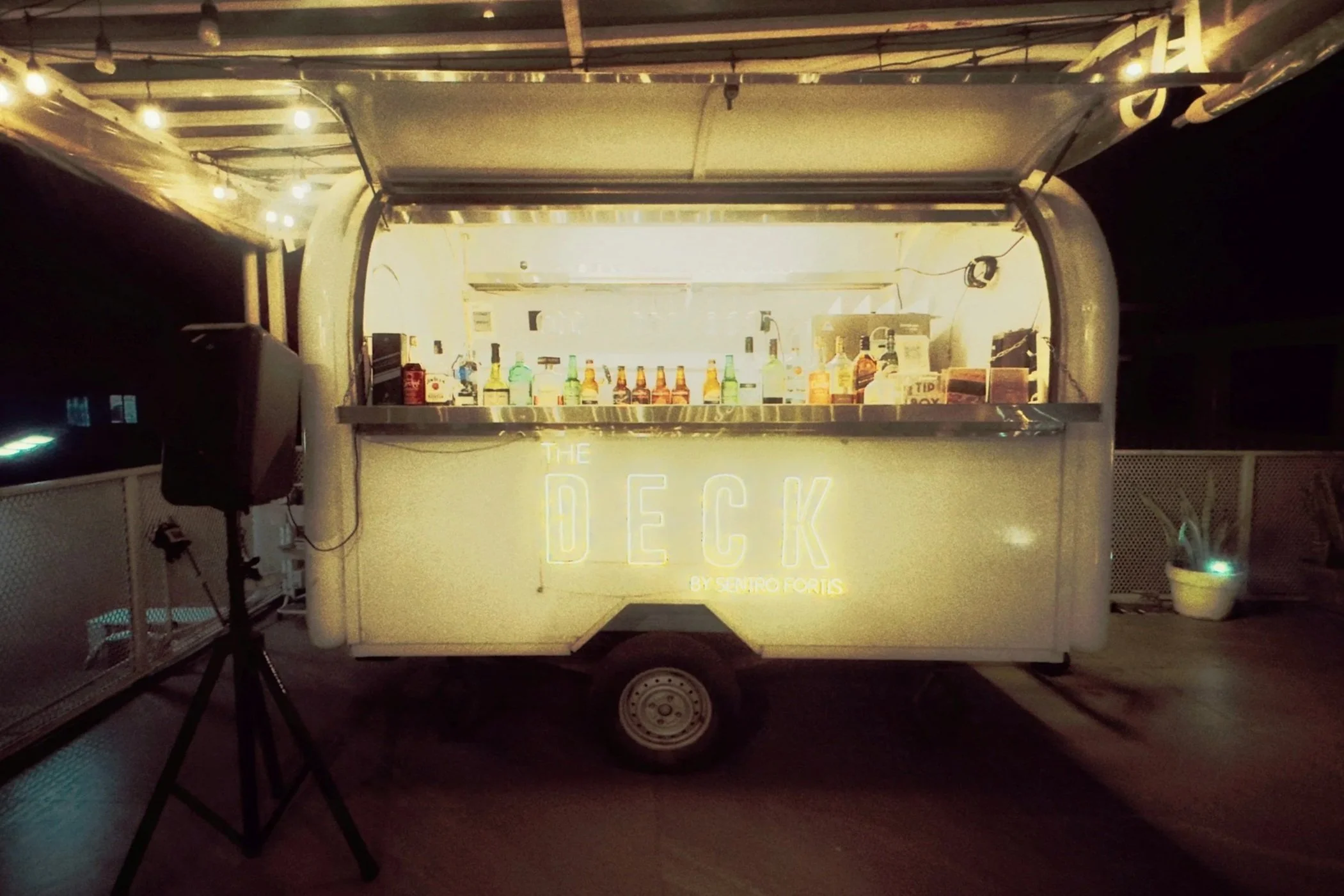 A mobile bar setup in a trailer with illuminated signage that reads 'The Deck by Sentro Fortis'. The trailer is stocked with various bottles of alcohol and mixers, and is set against a dark background with string lights overhead.