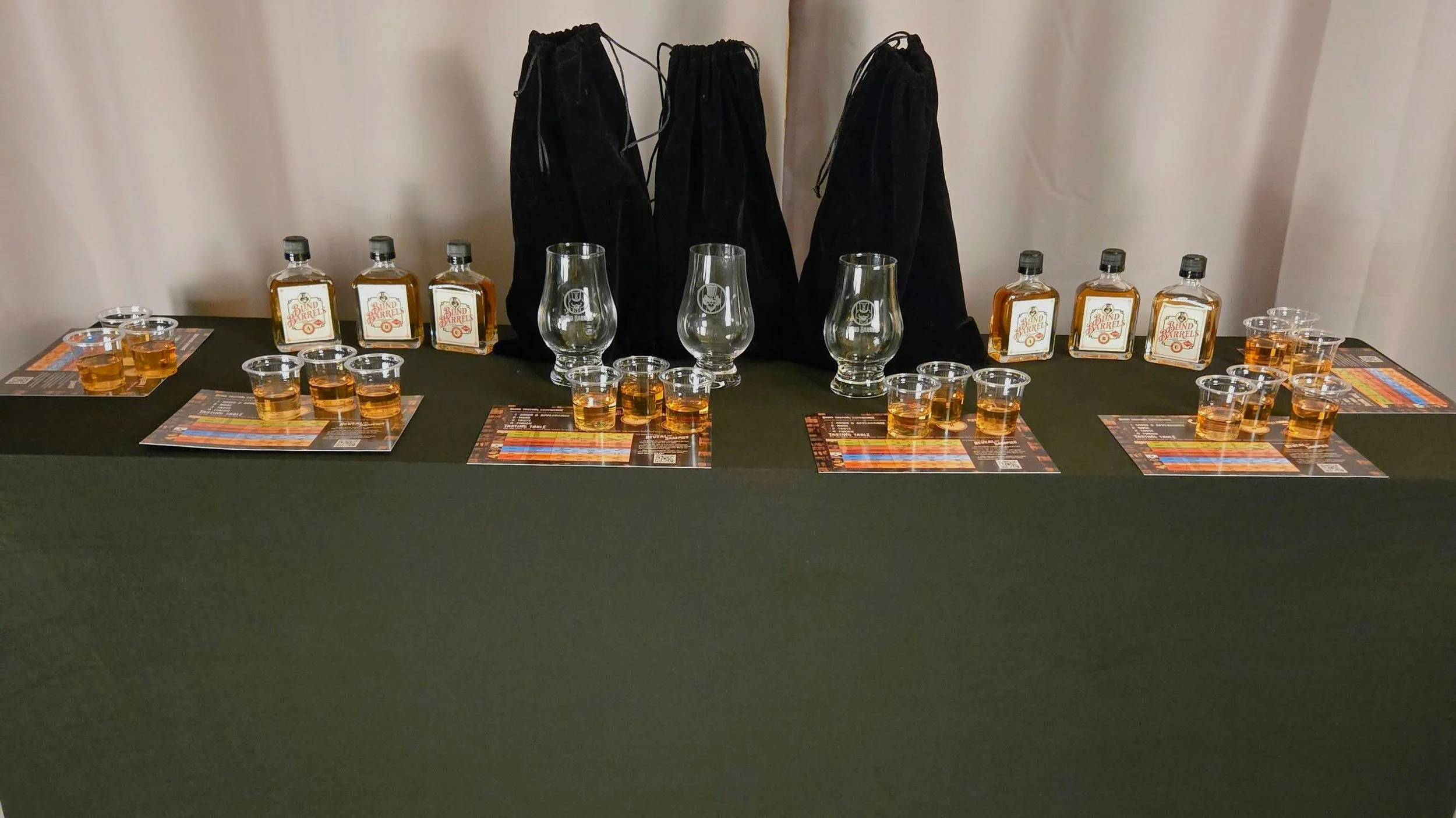 Display table with three bottles of bourbon, glasses with bourbon, taster glasses, educational mats, and black bag in the background.