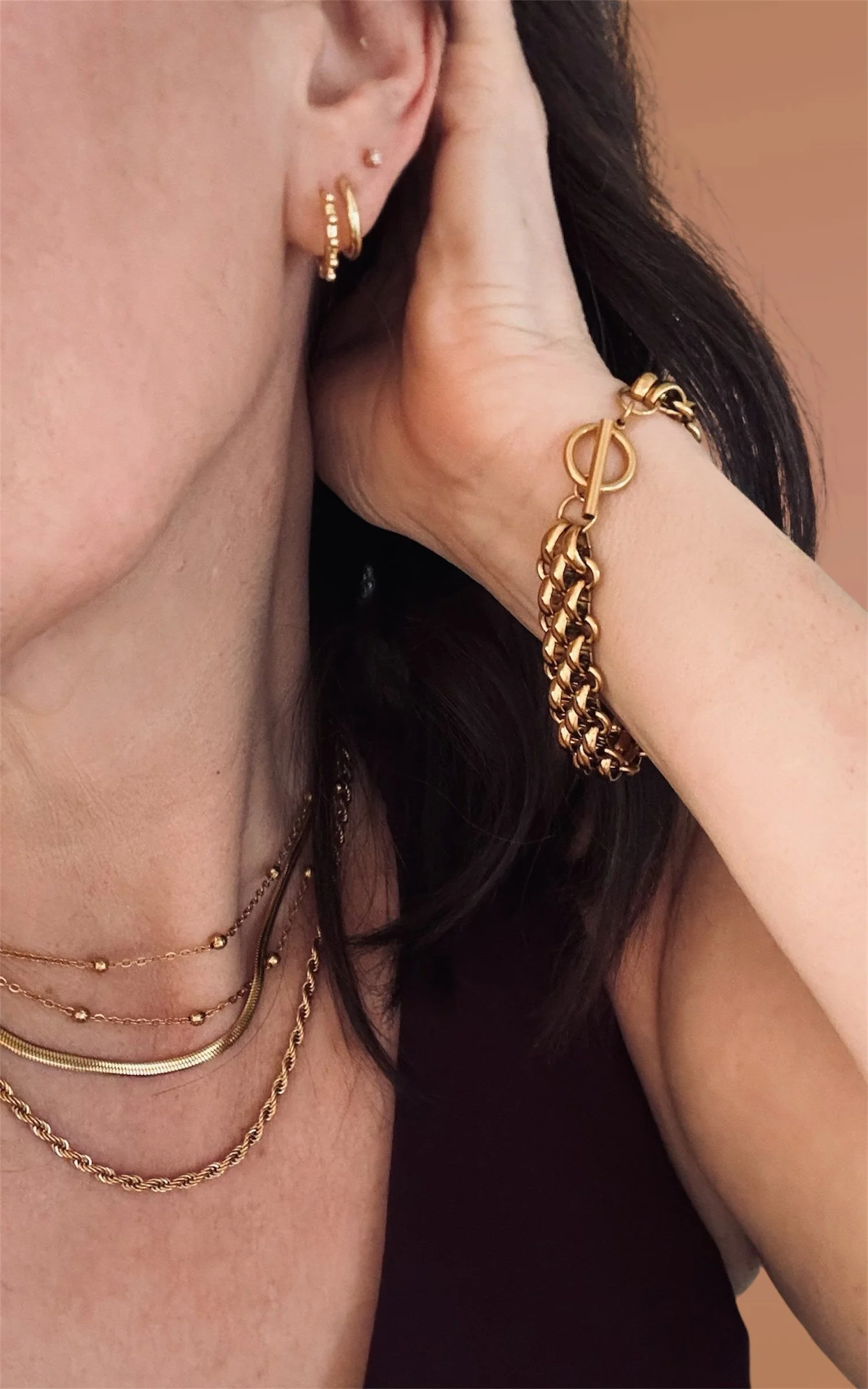 Close-up of a woman's jewelry, including three layered gold necklaces, a gold bracelet, and multiple gold earrings, with dark hair and a black top.