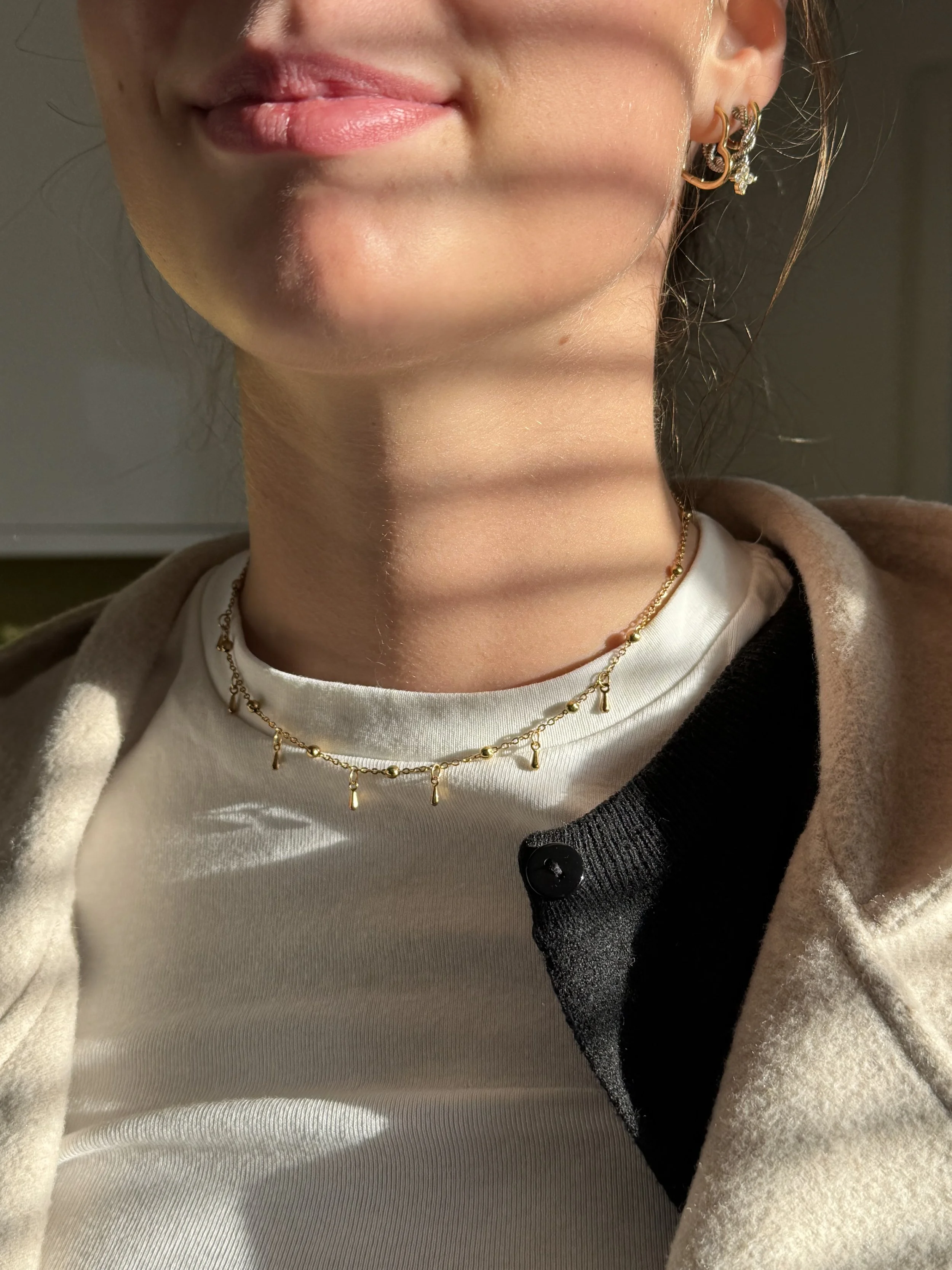 Close-up of a woman's neck and lower face, showing gold jewelry including a necklace and earrings, with sunlight creating striped shadows on her skin.