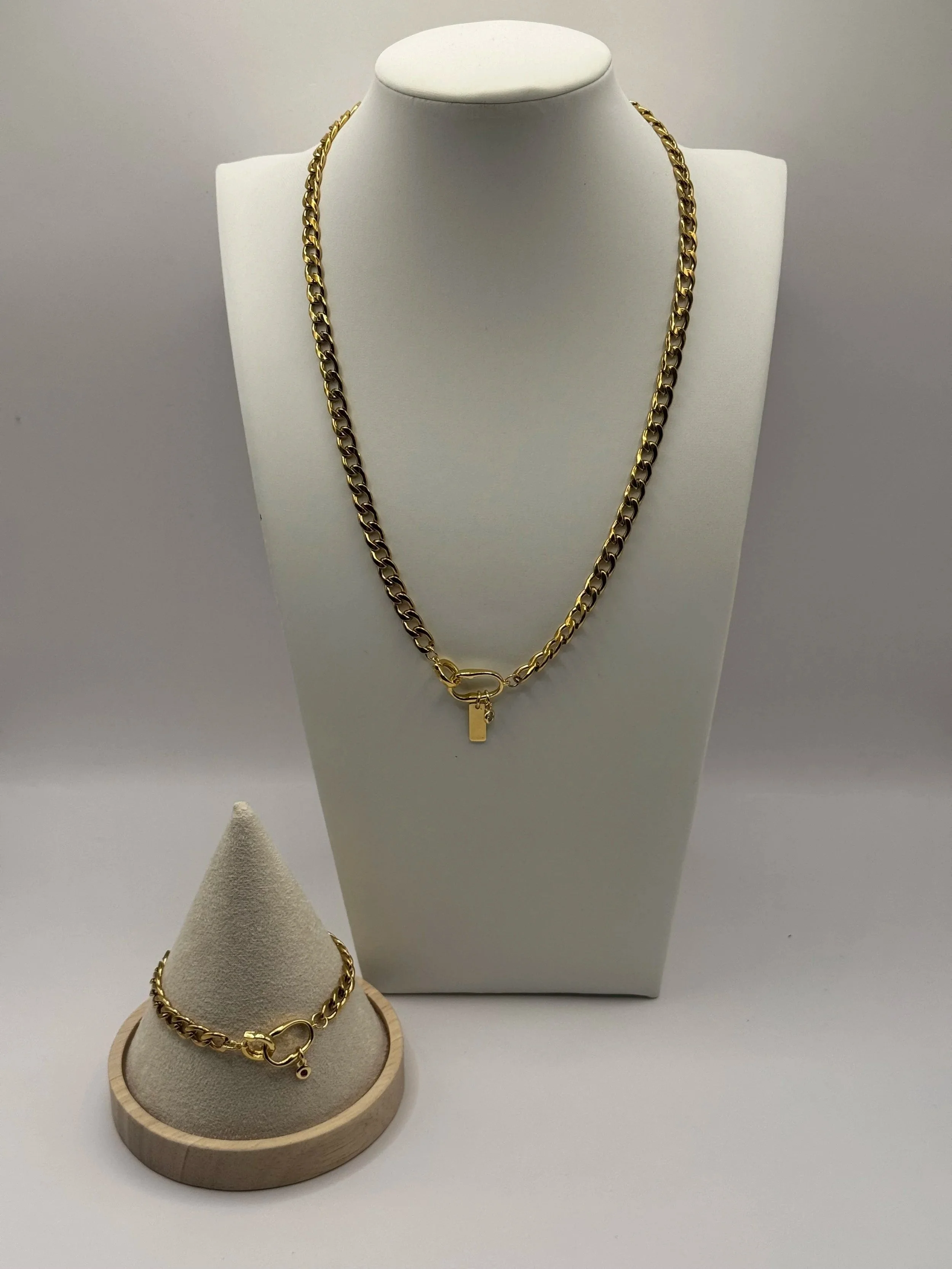 Gold chain necklace with a small padlock charm and a bracelet with a similar design, displayed on a white jewelry stand and a beige jewelry cone