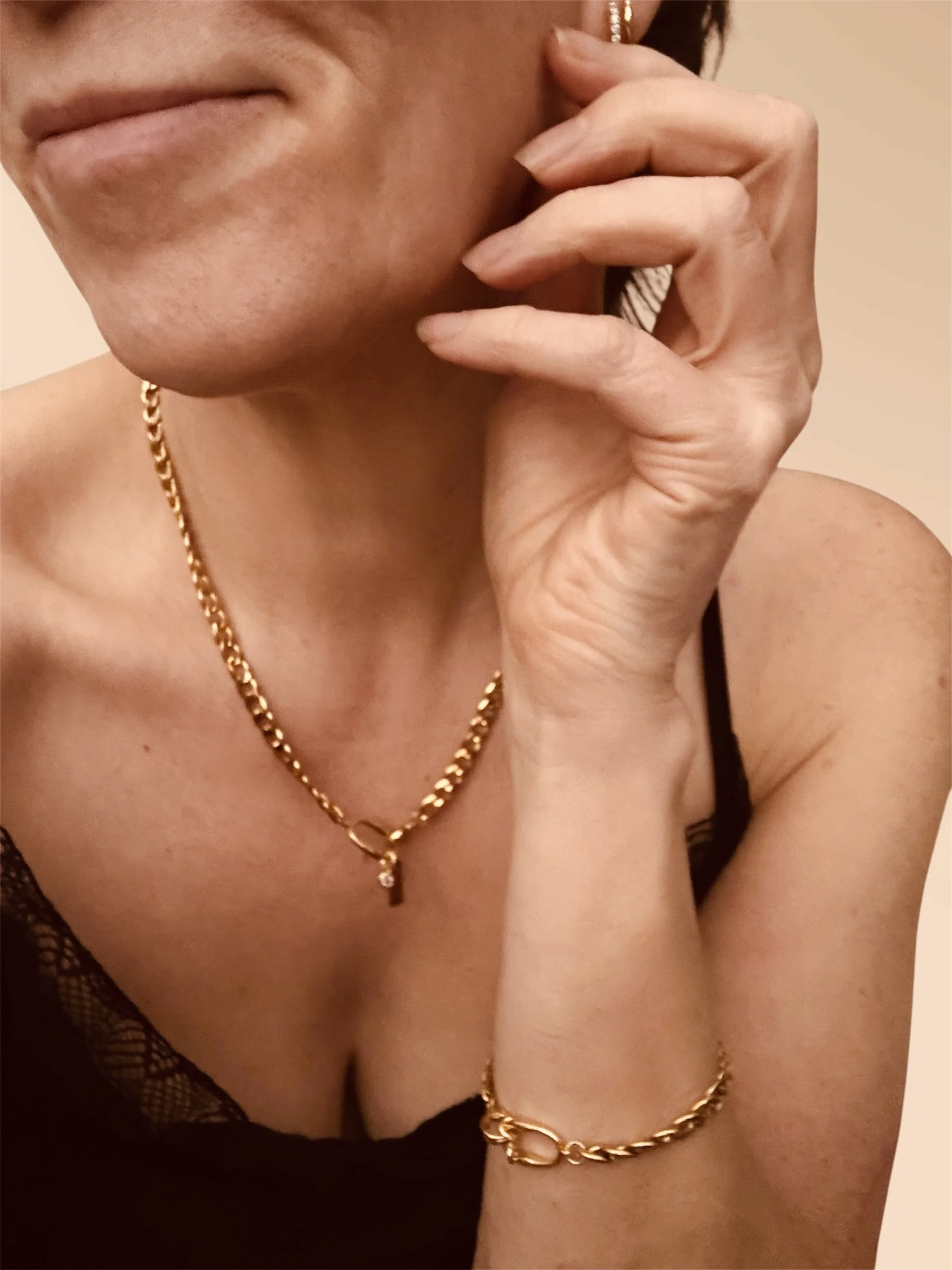 Close-up of a woman's face and hand, showing gold jewelry including a necklace and bracelet, and wearing a black spaghetti strap top.