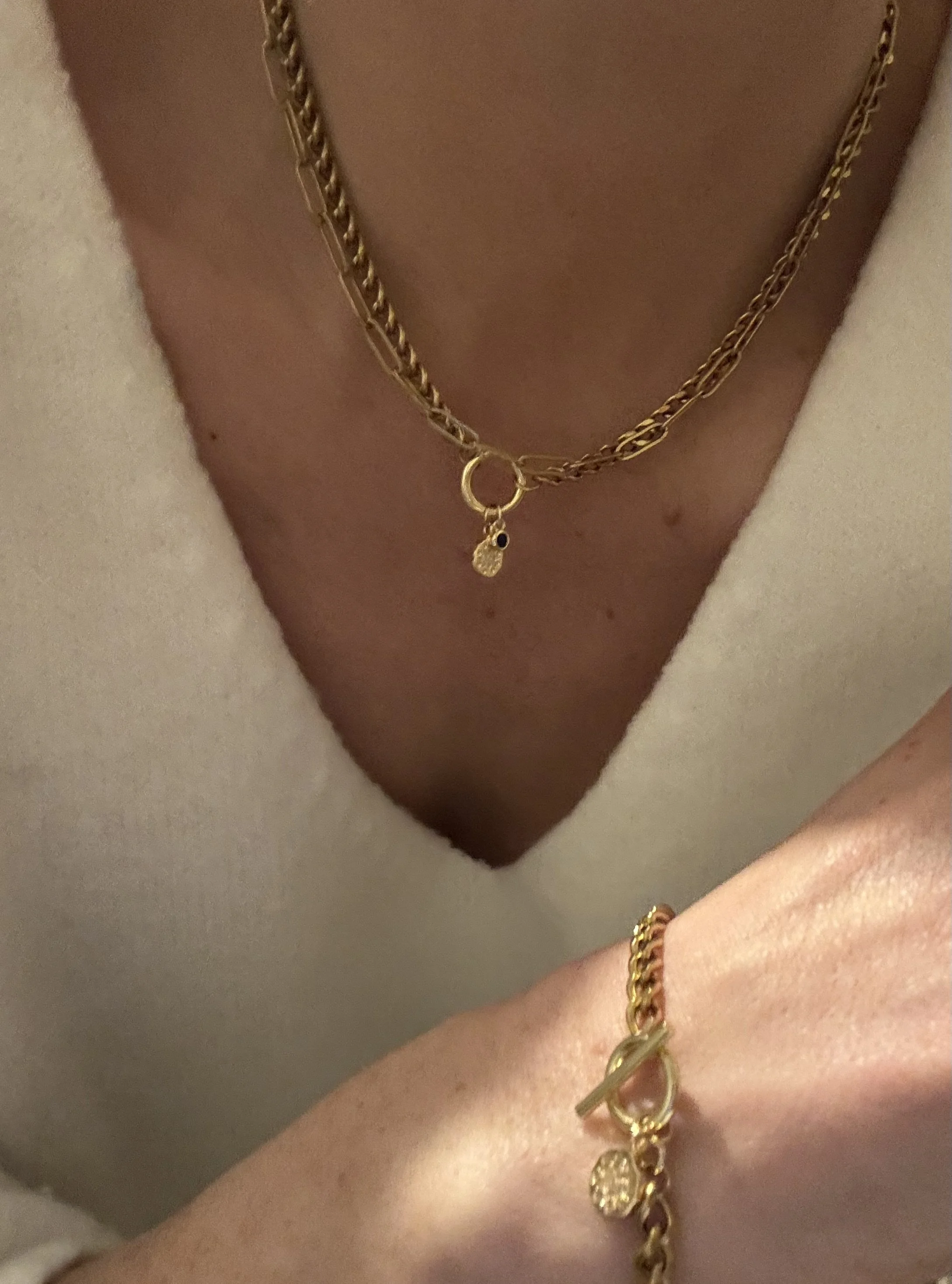 Close-up of a person's neck and wrist showing gold jewelry, including a layered gold chain necklace with a small pendant, and a gold bracelet with a clasp and charms.
