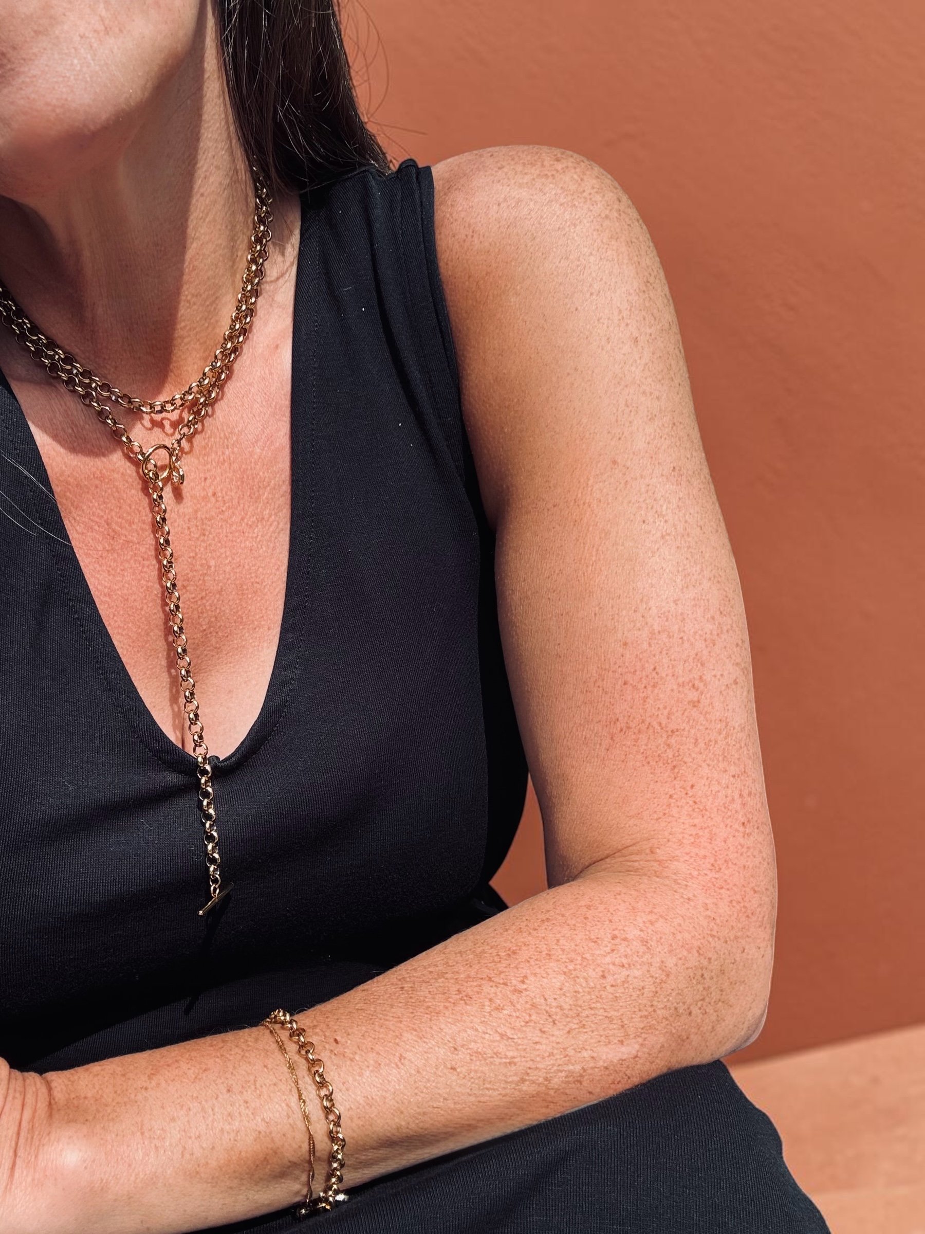 Close-up of a woman wearing layered gold chains and a gold bracelet, dressed in a red top, with pink flowers in the background.