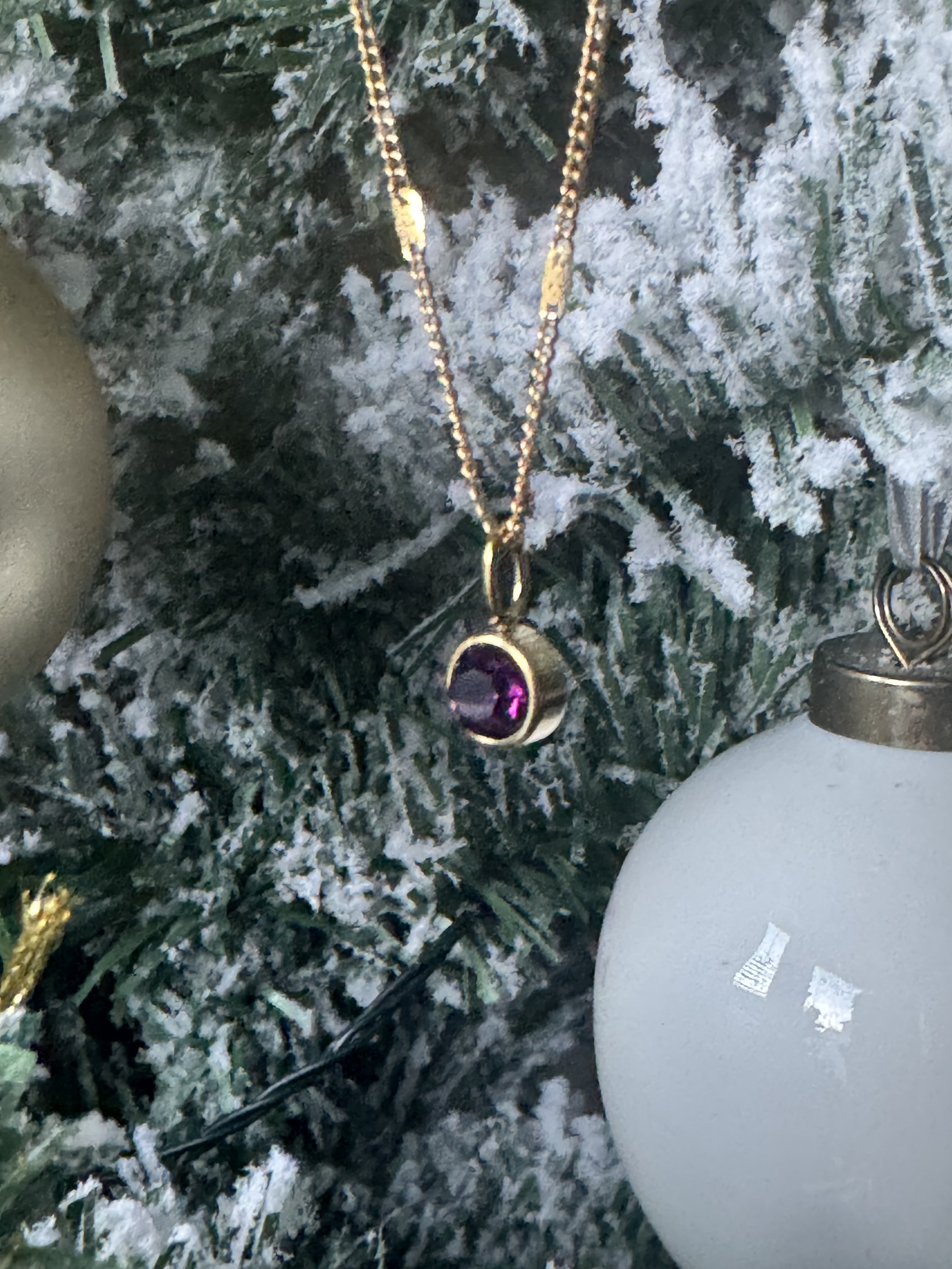 A close-up of a Christmas tree decorated with snow-like frosting, a gold chain with a purple gemstone pendant, and white ornaments.