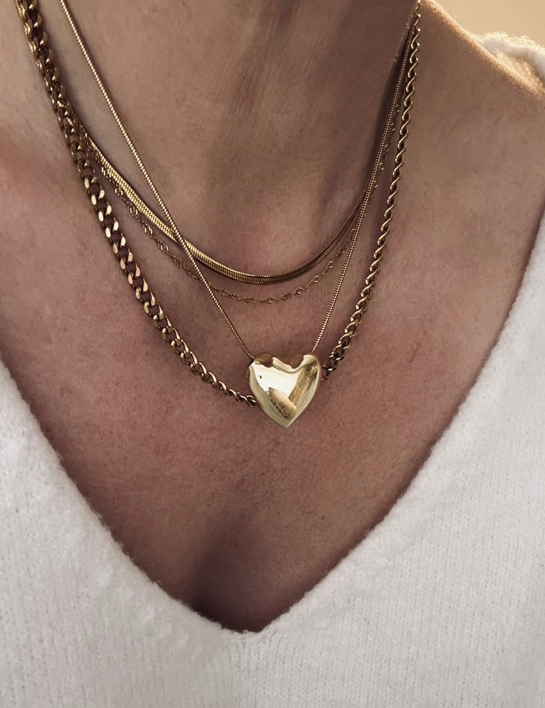 Multiple layered gold necklaces, including one with a heart-shaped pendant, worn over a white V-neck top.