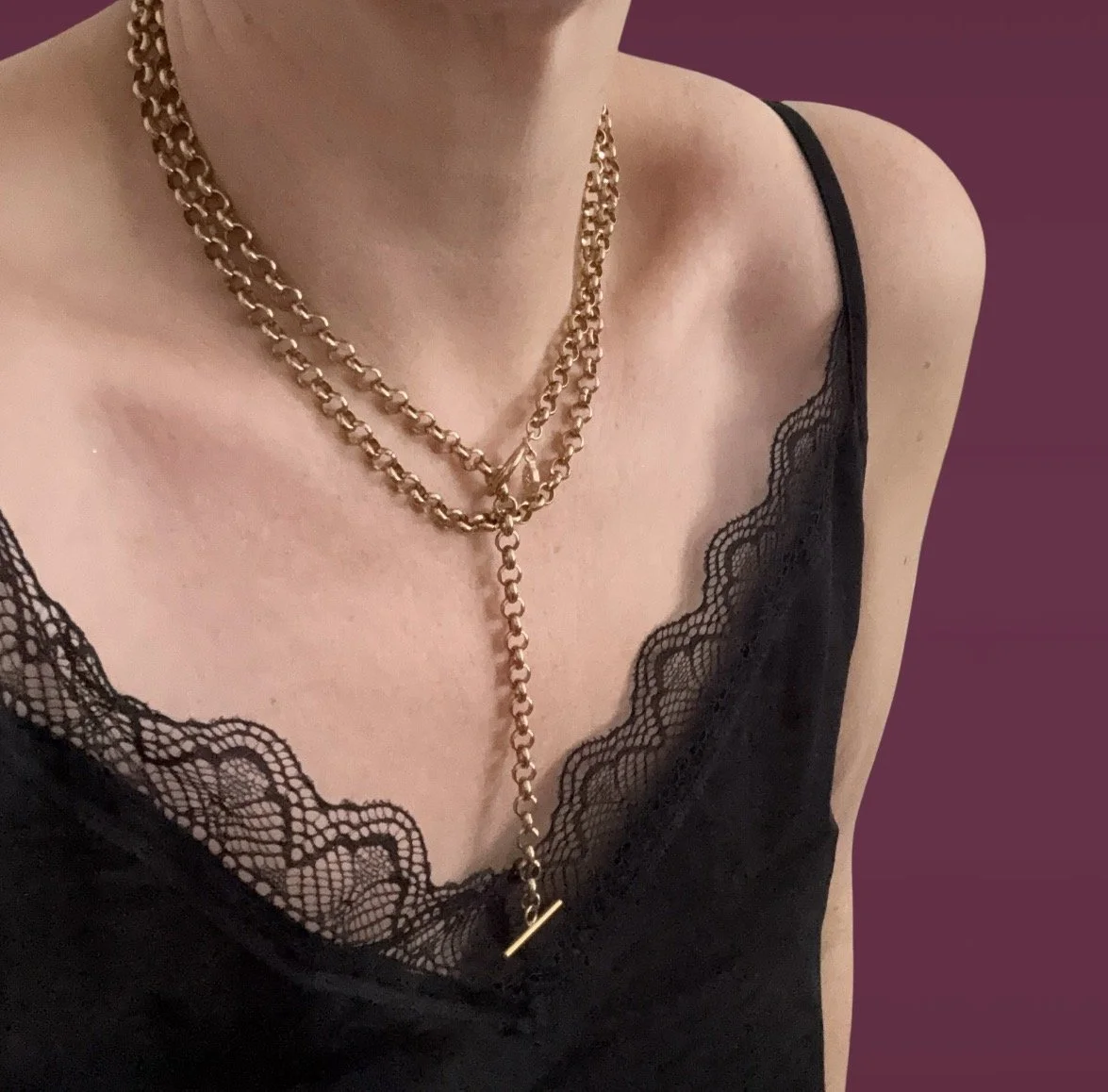 Close-up of a woman's chest showing layered gold chain necklaces against a black lace slip top with spaghetti straps and a purple background.