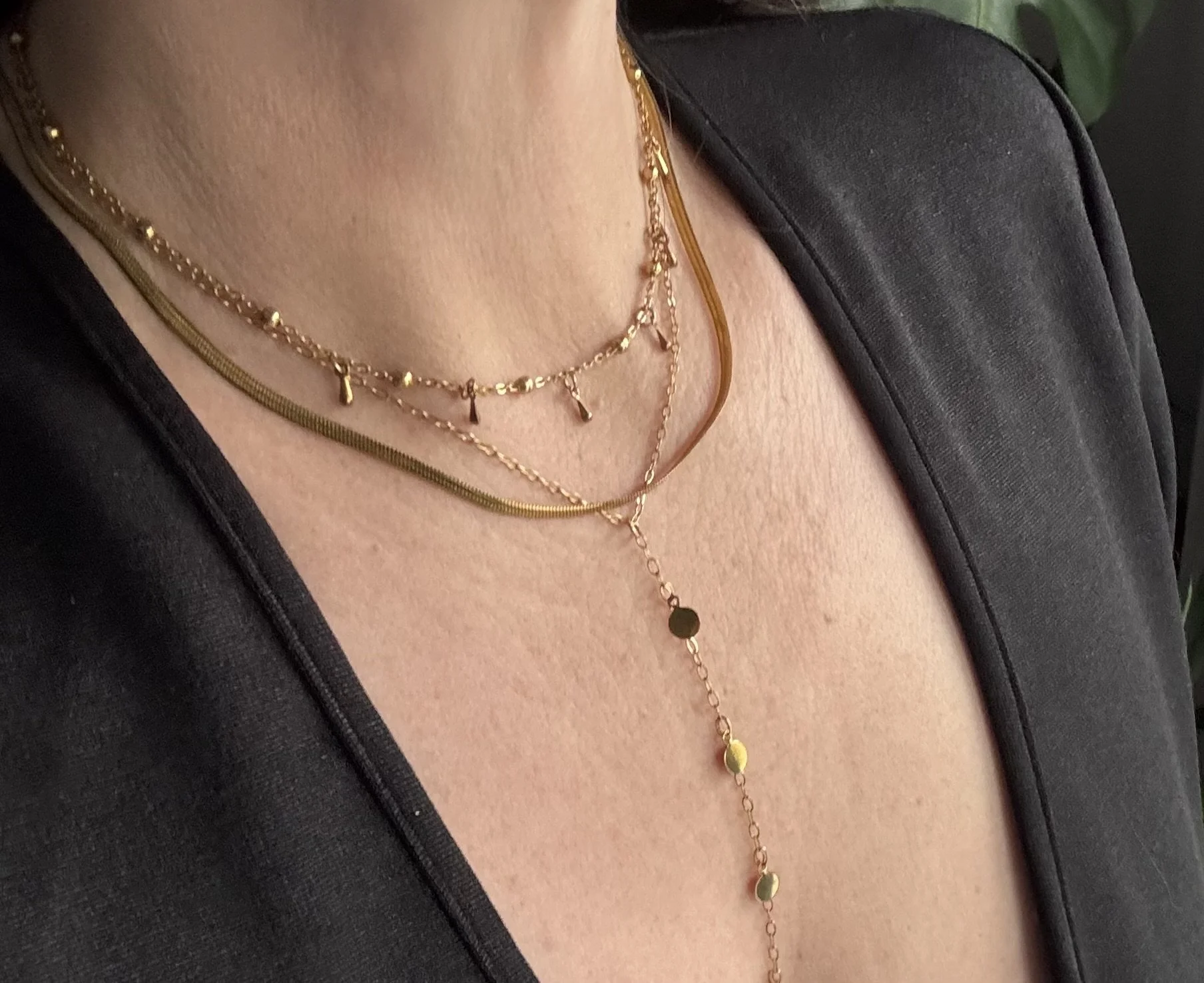 Close-up of a woman's neck wearing three gold layered necklaces with small charms, over a black top or jacket.
