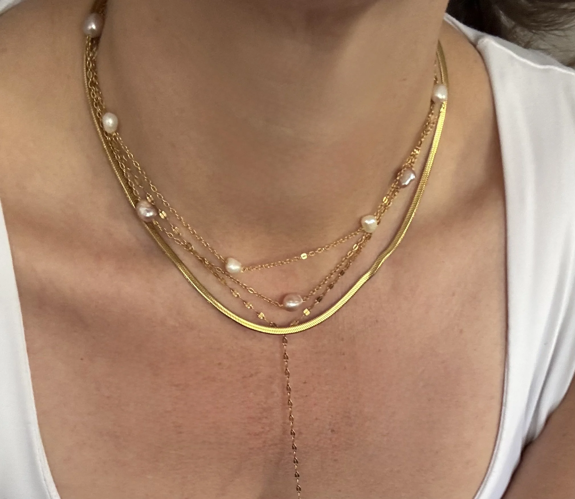 Close-up of a person's neck and upper chest wearing three layered gold necklaces with pearls, against a white top.