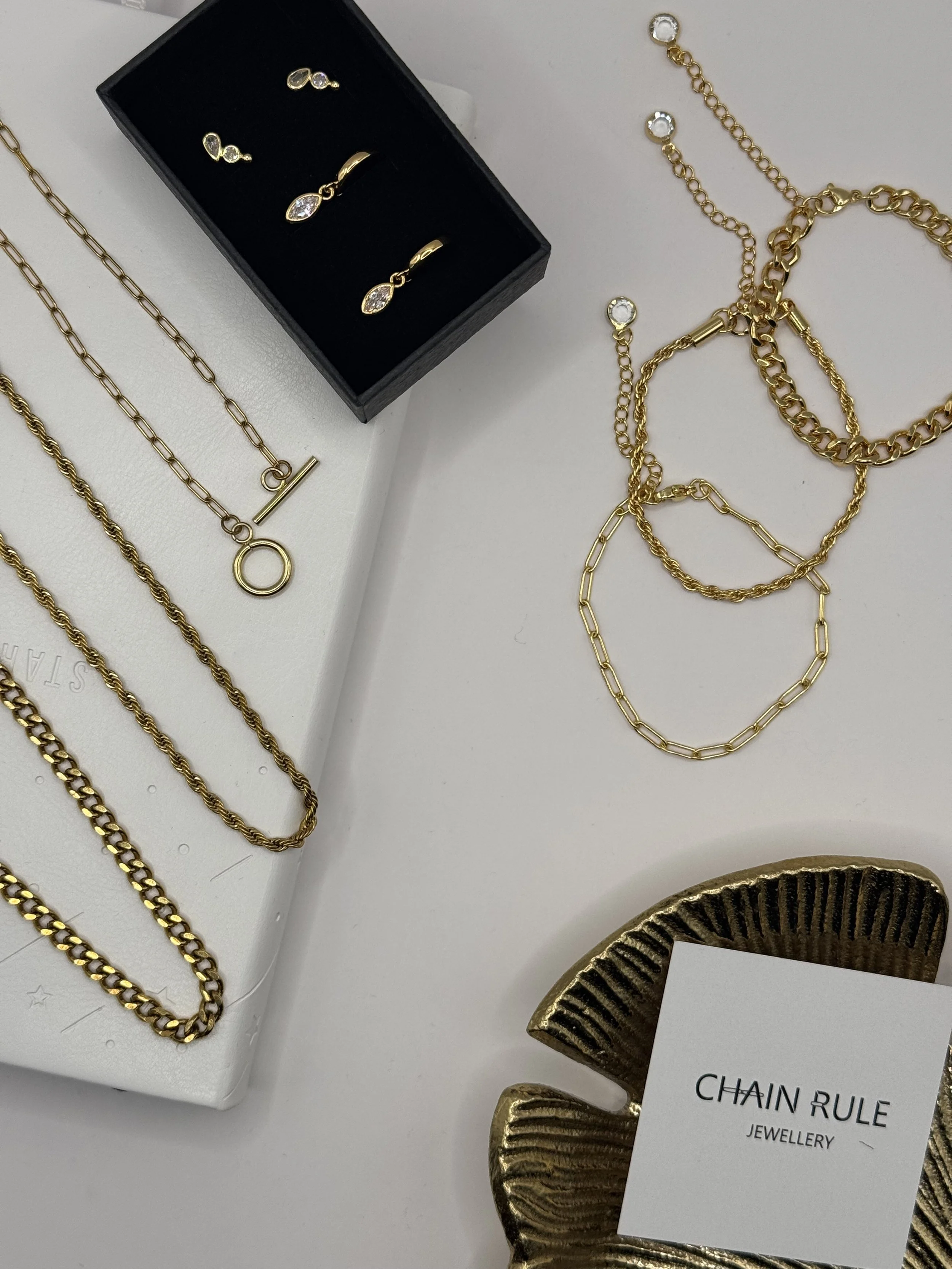 Gold jewelry set including necklaces, earrings, a ring, and bracelets displayed on a white surface with a jewelry business card that reads "CHAIN RULE JEWELLERY."