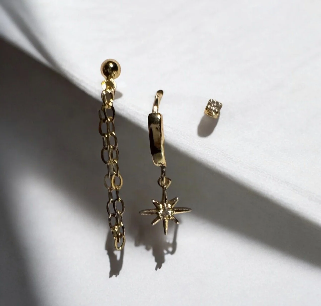 Two gold earrings and a small gold stud earring on a white surface with shadows.