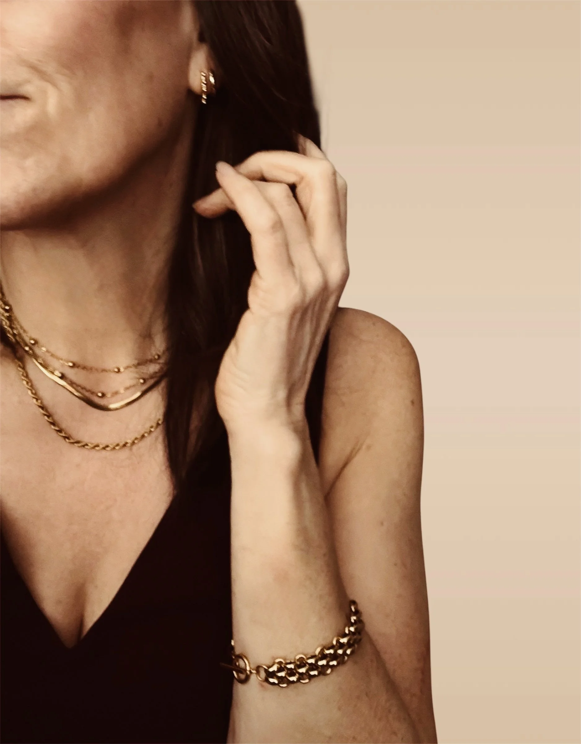 Close-up of a woman wearing gold jewelry, including layered necklaces, earrings, and a bracelet, with her hand near her face and a black top.