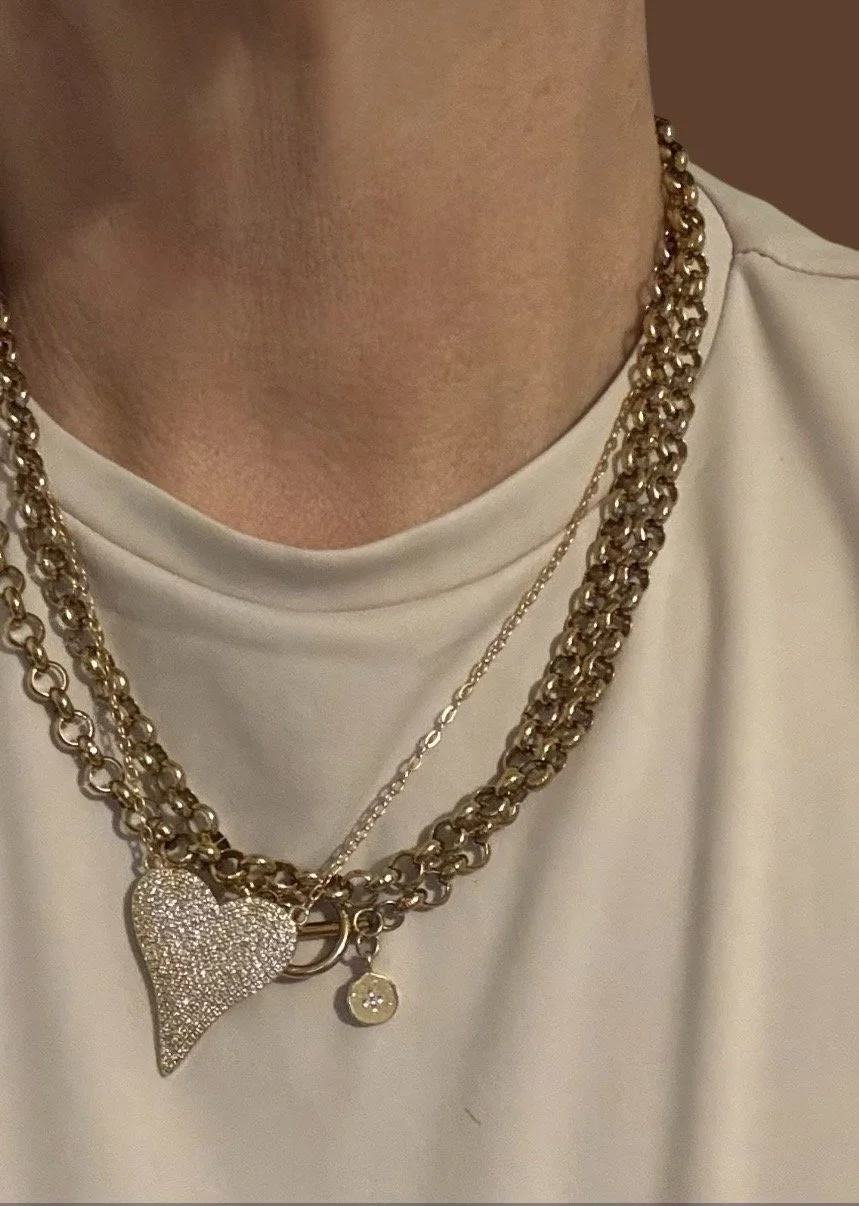 Close-up of a person's neck and chest showing layered gold necklaces, including a heart-shaped pendant covered in small stones, a small circular pendant, and a thin chain, against a white shirt.